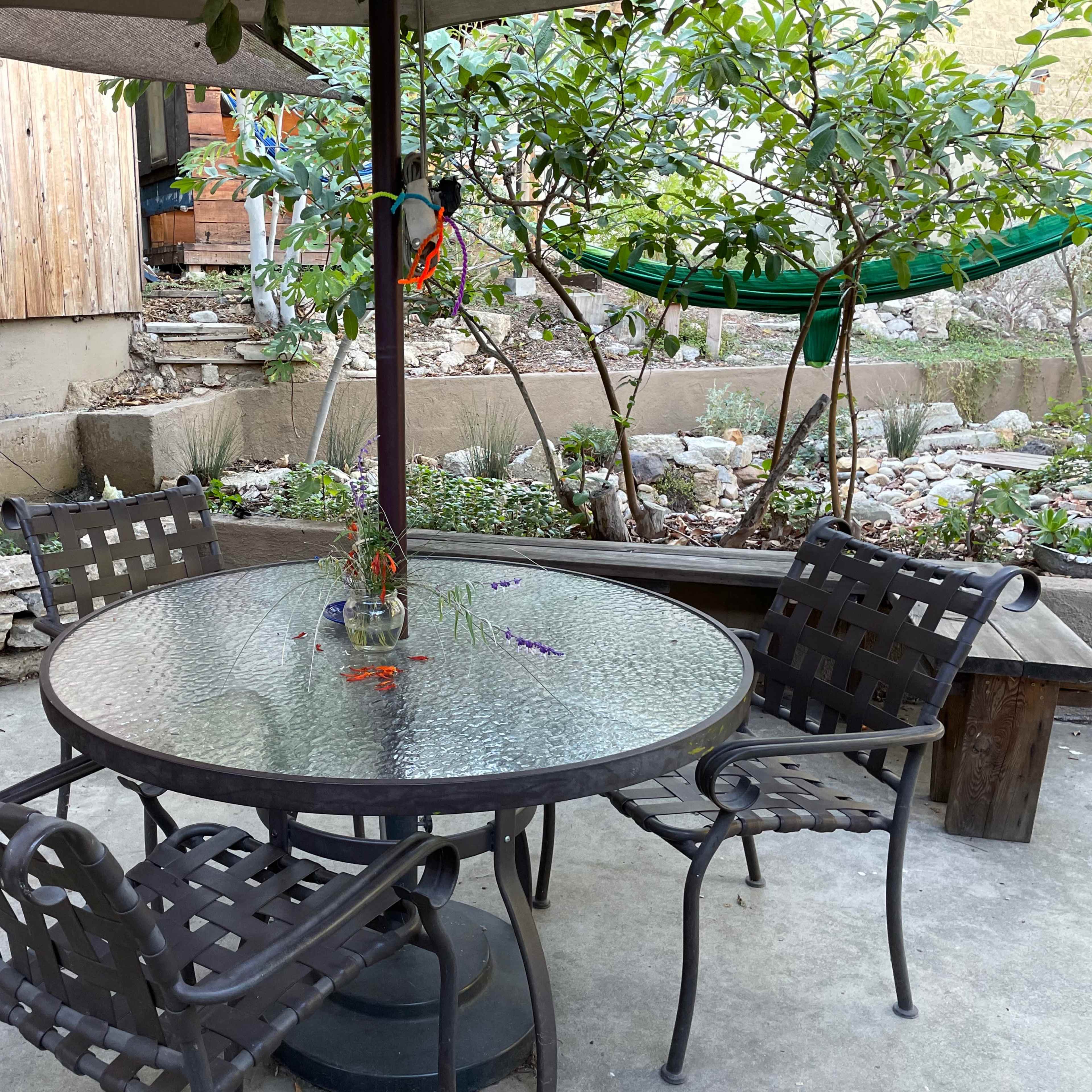 A circular glass-top table is surrounded by four metal chairs under a shaded pergola in a backyard setting with greenery.