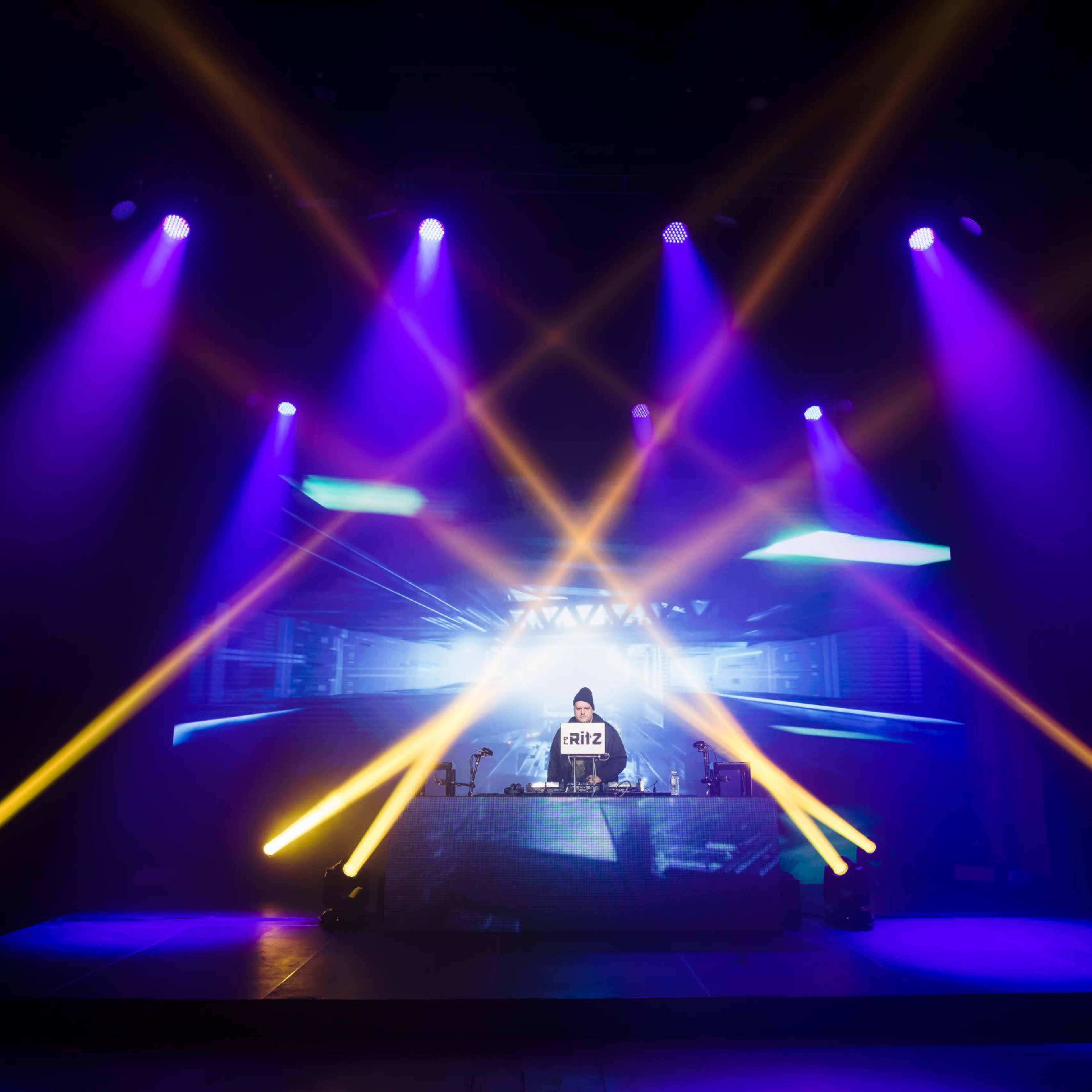 A DJ performs on stage with vibrant lighting effects and a large visual display behind them.