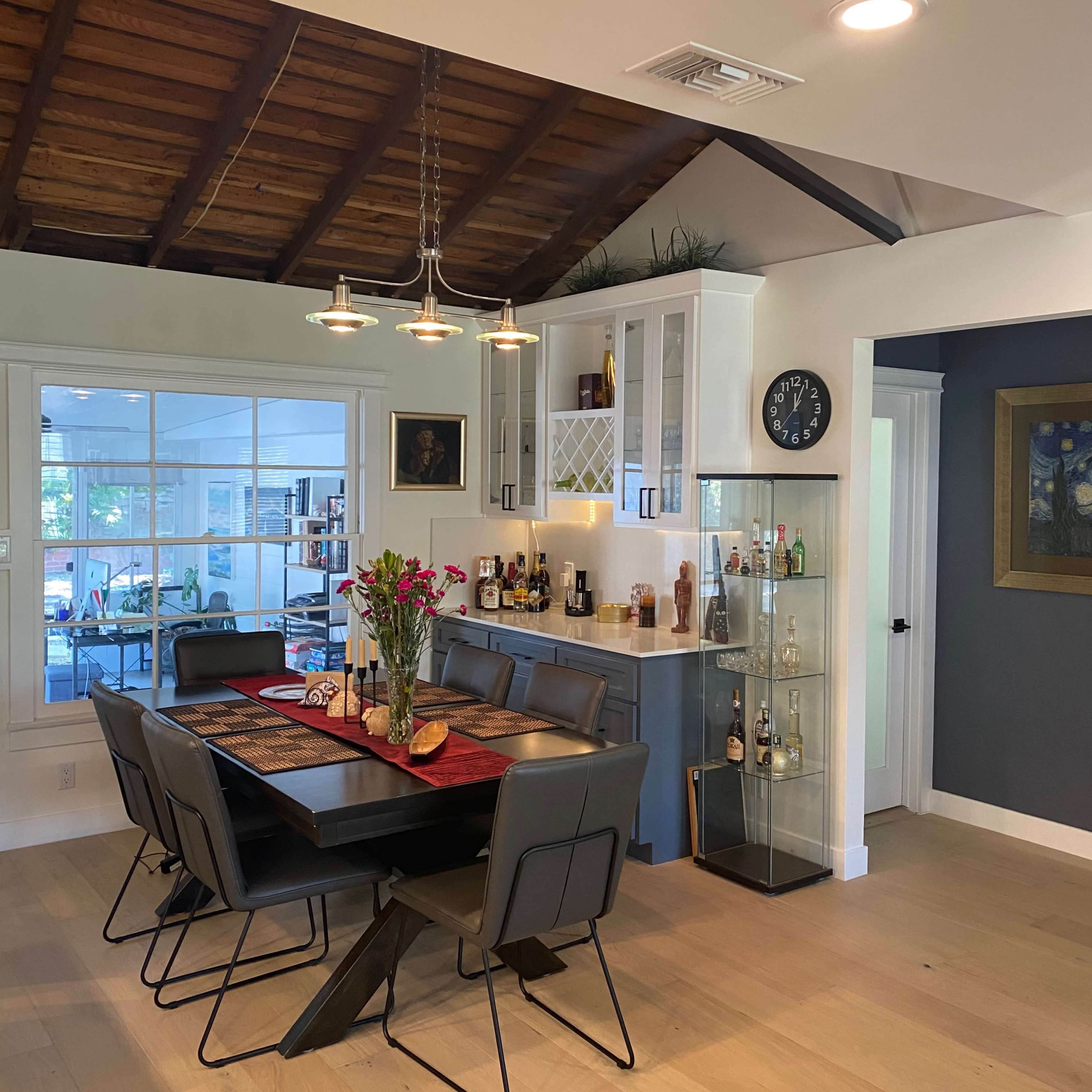 A dining area features a wooden table with black chairs, surrounded by a kitchen with modern cabinetry and a display cabinet for bottles.