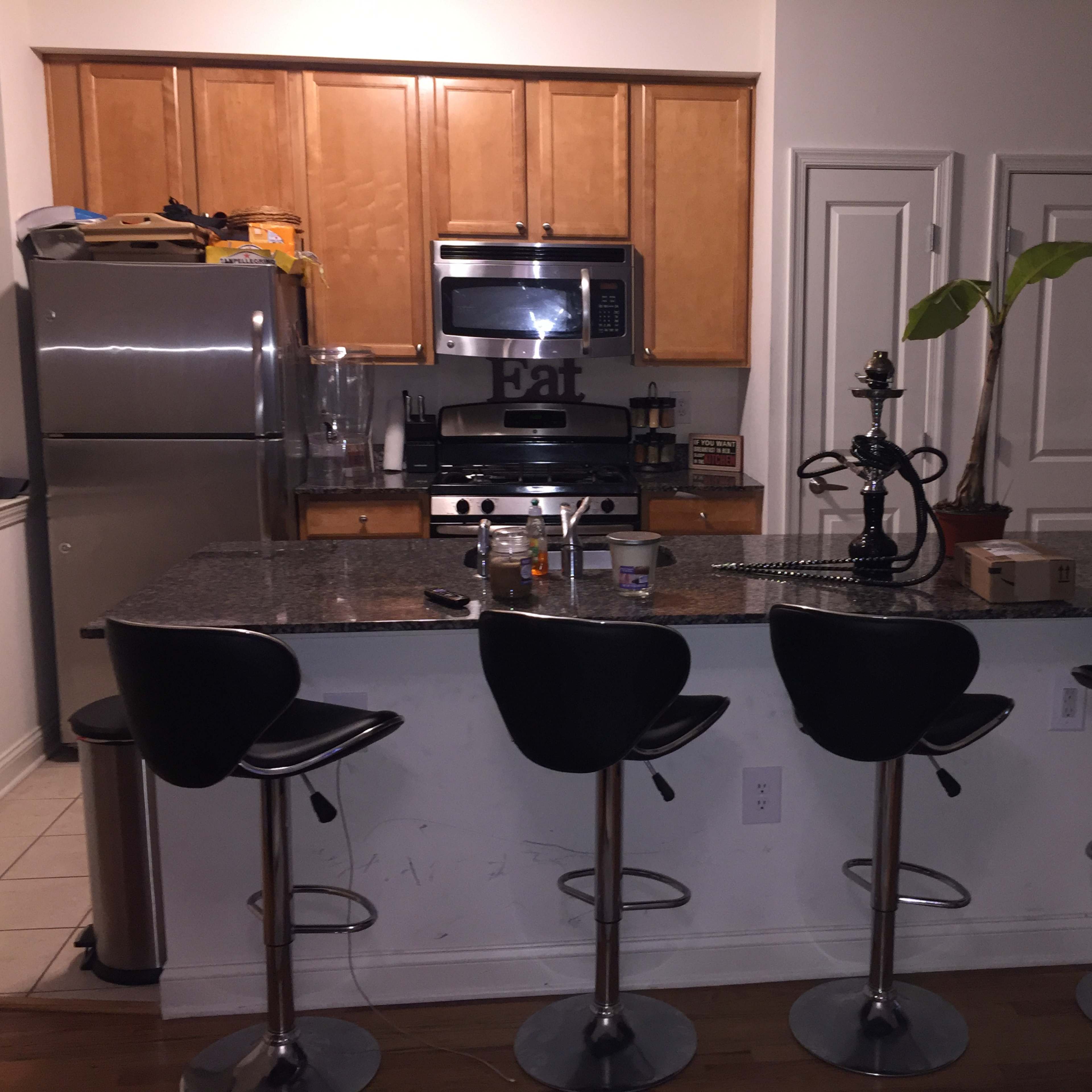 A modern kitchen with a bar area featuring four black stools, a stainless steel refrigerator, an oven, and a countertop with various items.