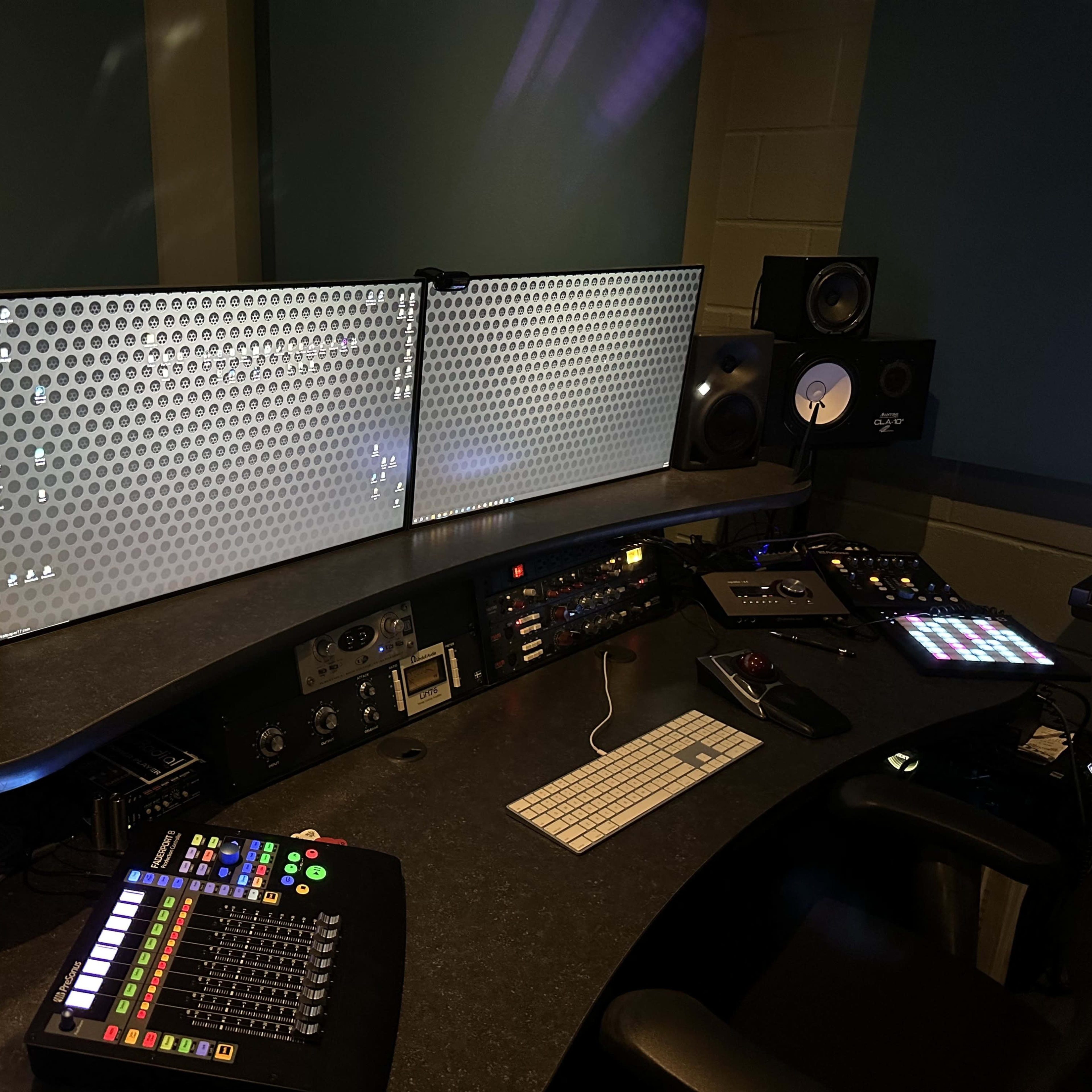 A music production studio featuring dual monitors, a mixing console, various audio equipment, and speakers arranged on a U-shaped desk.