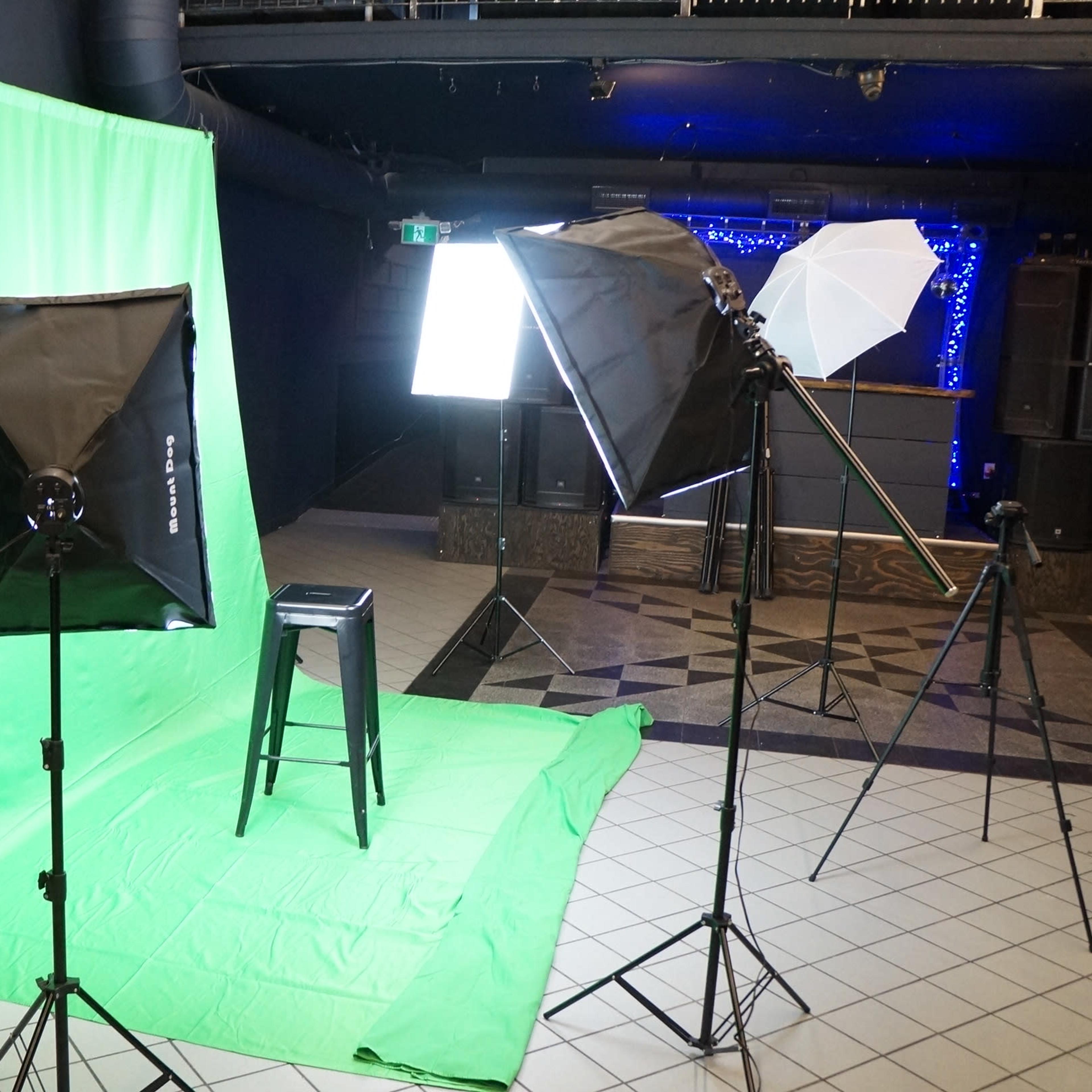 A photography setup featuring a green backdrop, a black stool, and several studio lights arranged on tripods in a spacious room with a patterned floor.