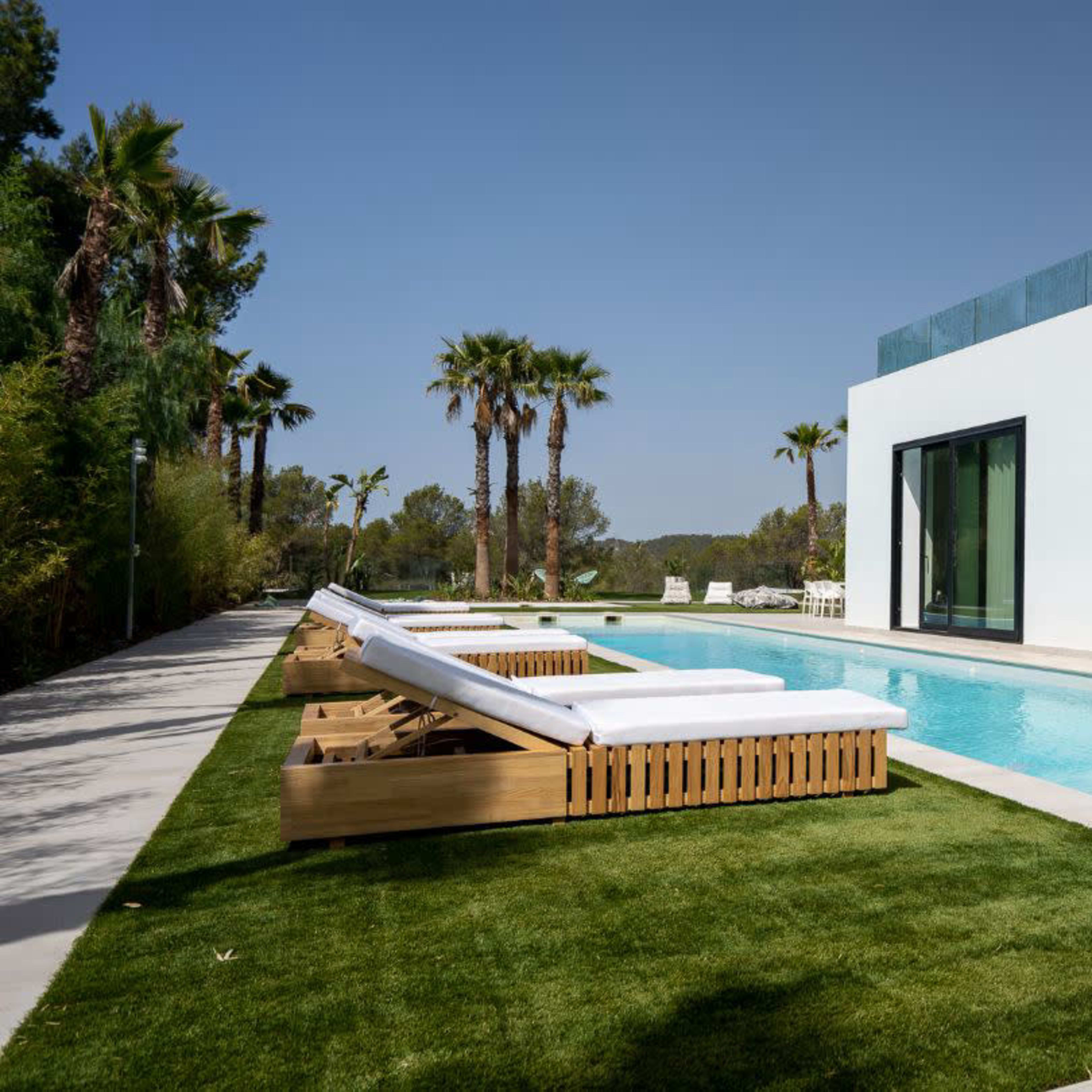 A modern villa features a sleek swimming pool surrounded by palm trees and lounge chairs on a grassy area.
