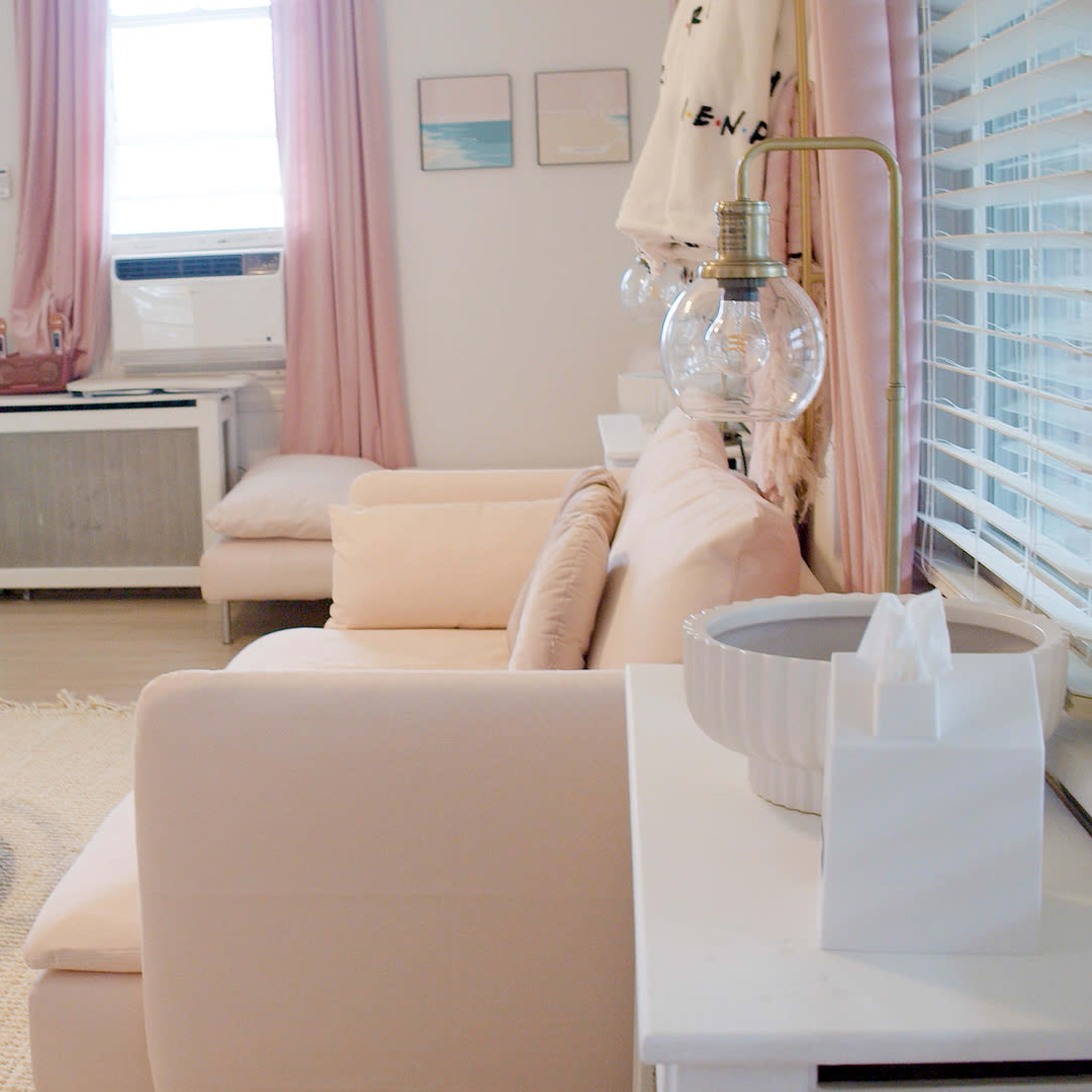 A cozy living room with a light-colored sofa, a circular glass coffee table, and soft pink curtains by a window.