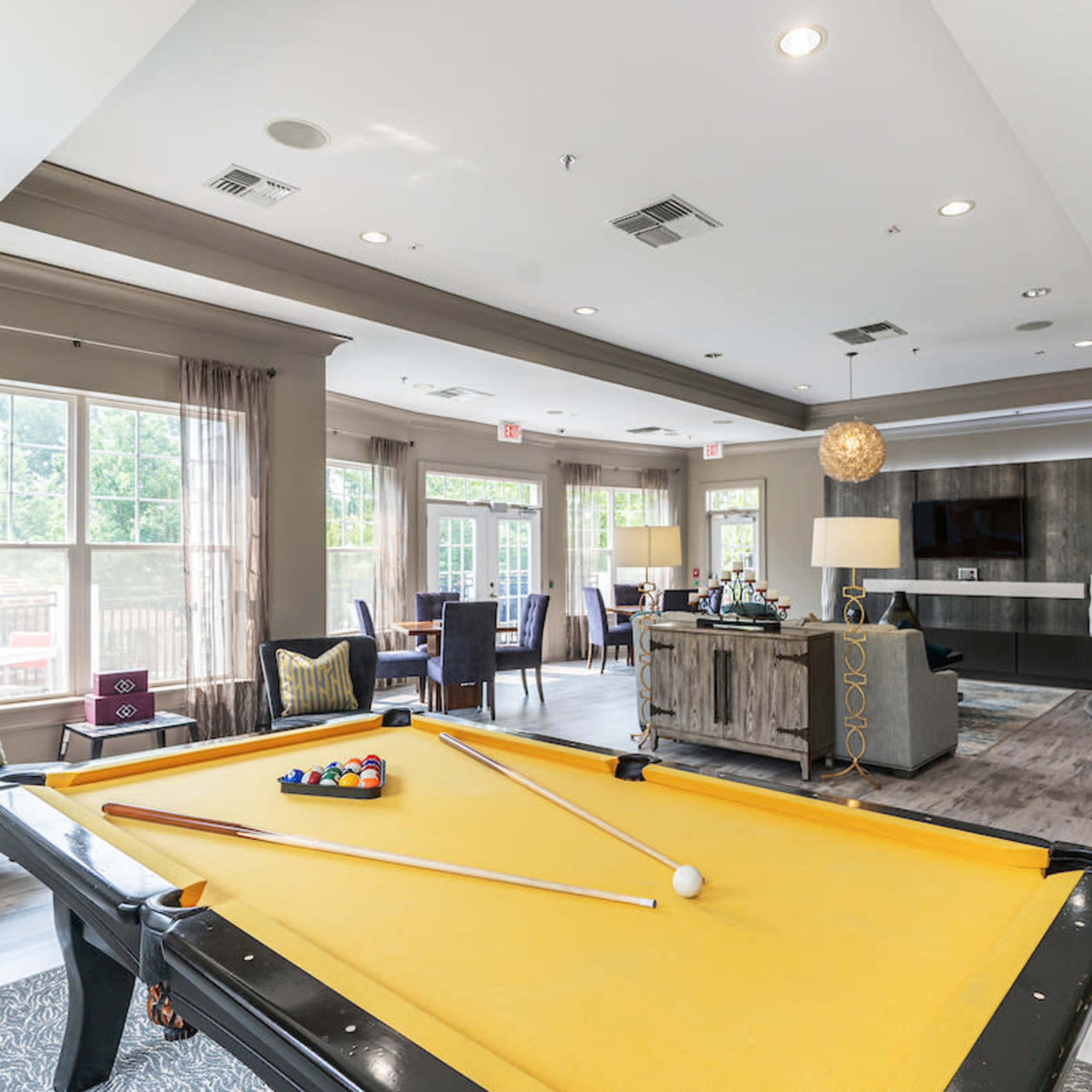 A well-lit recreational room featuring a yellow pool table, seating areas, and large windows with views outside.