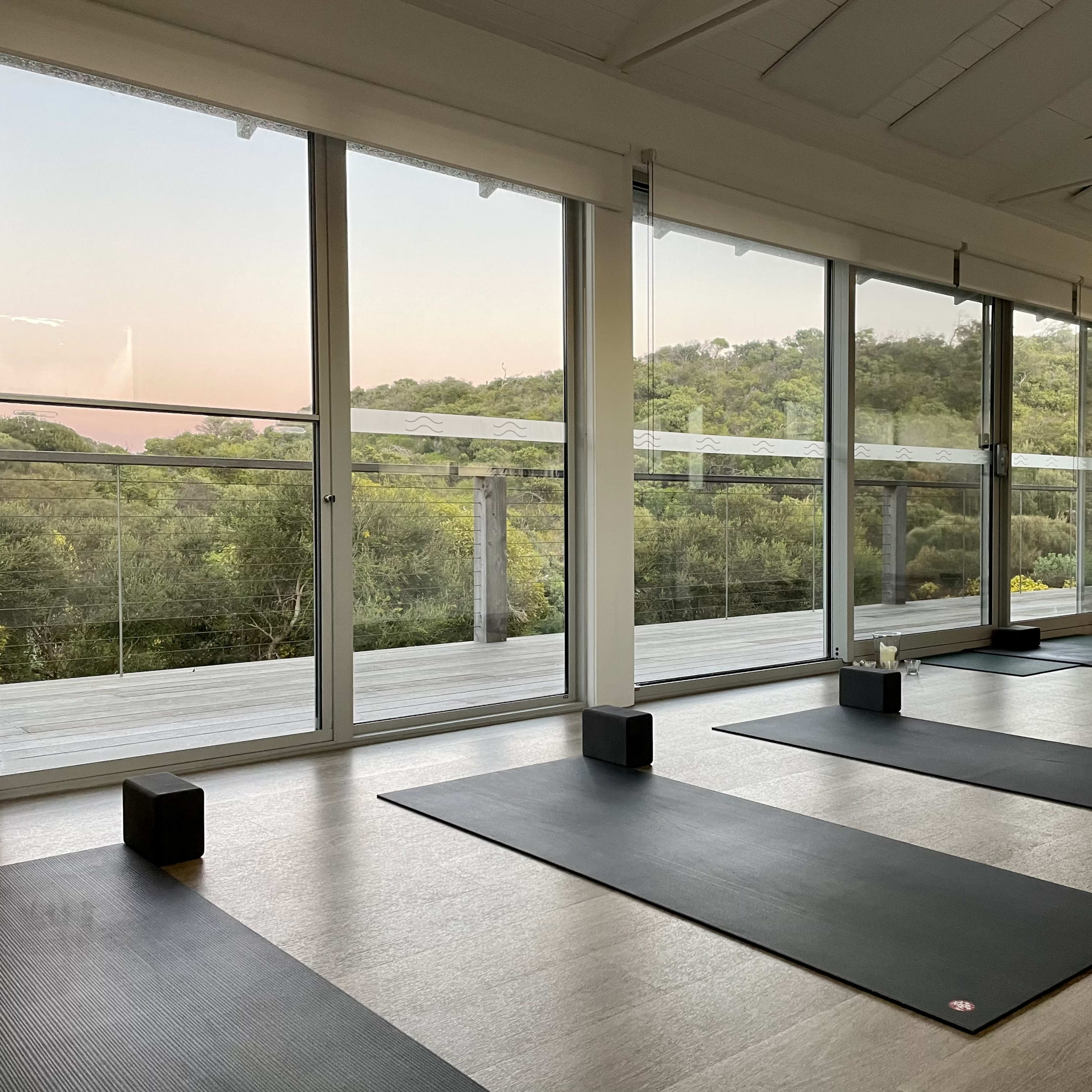 A yoga studio with wooden floors and yoga mats arranged neatly beside large windows that overlook a green hillside.