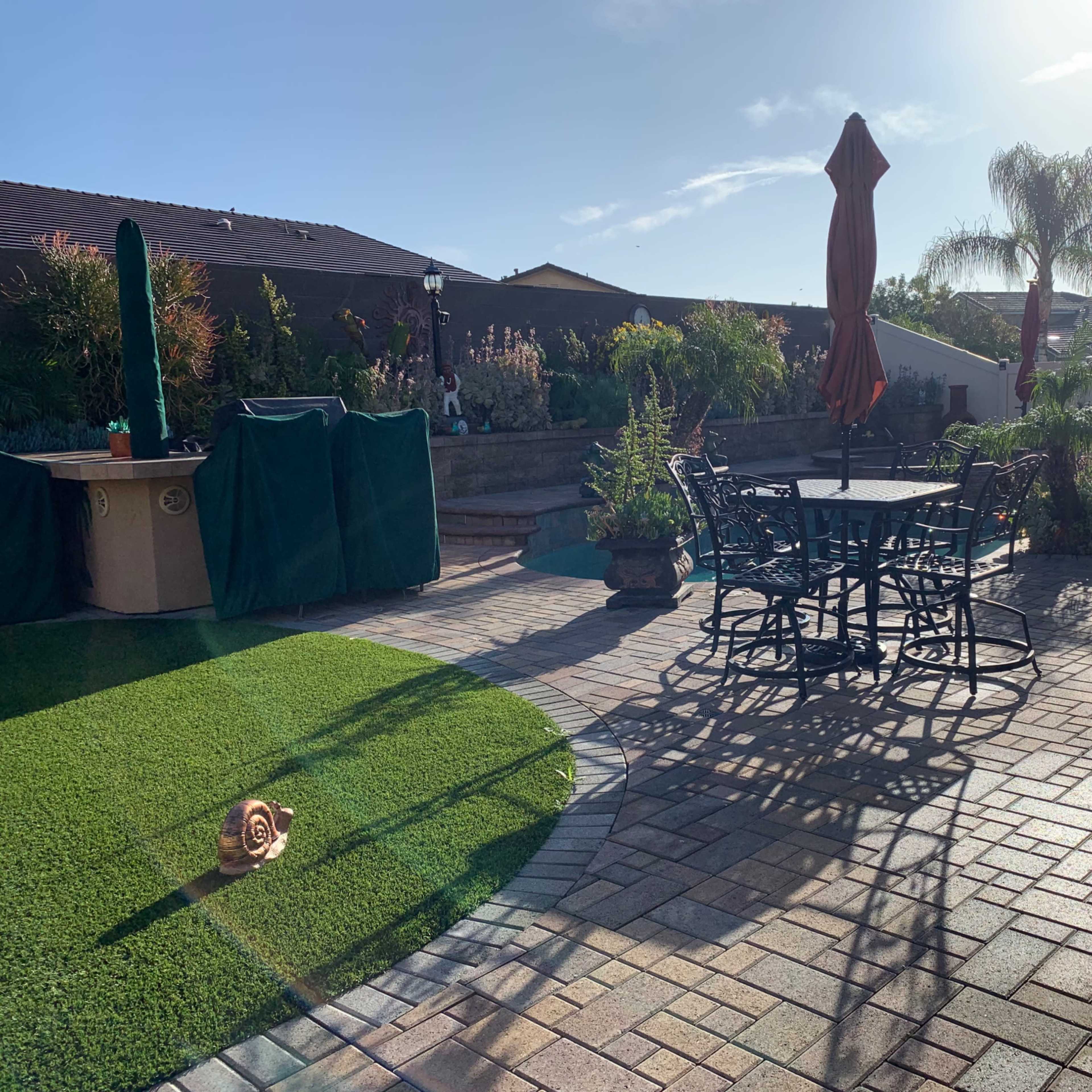 A landscaped backyard features a patio with a wrought-iron table and chairs, surrounded by potted plants and a small artificial grass area.
