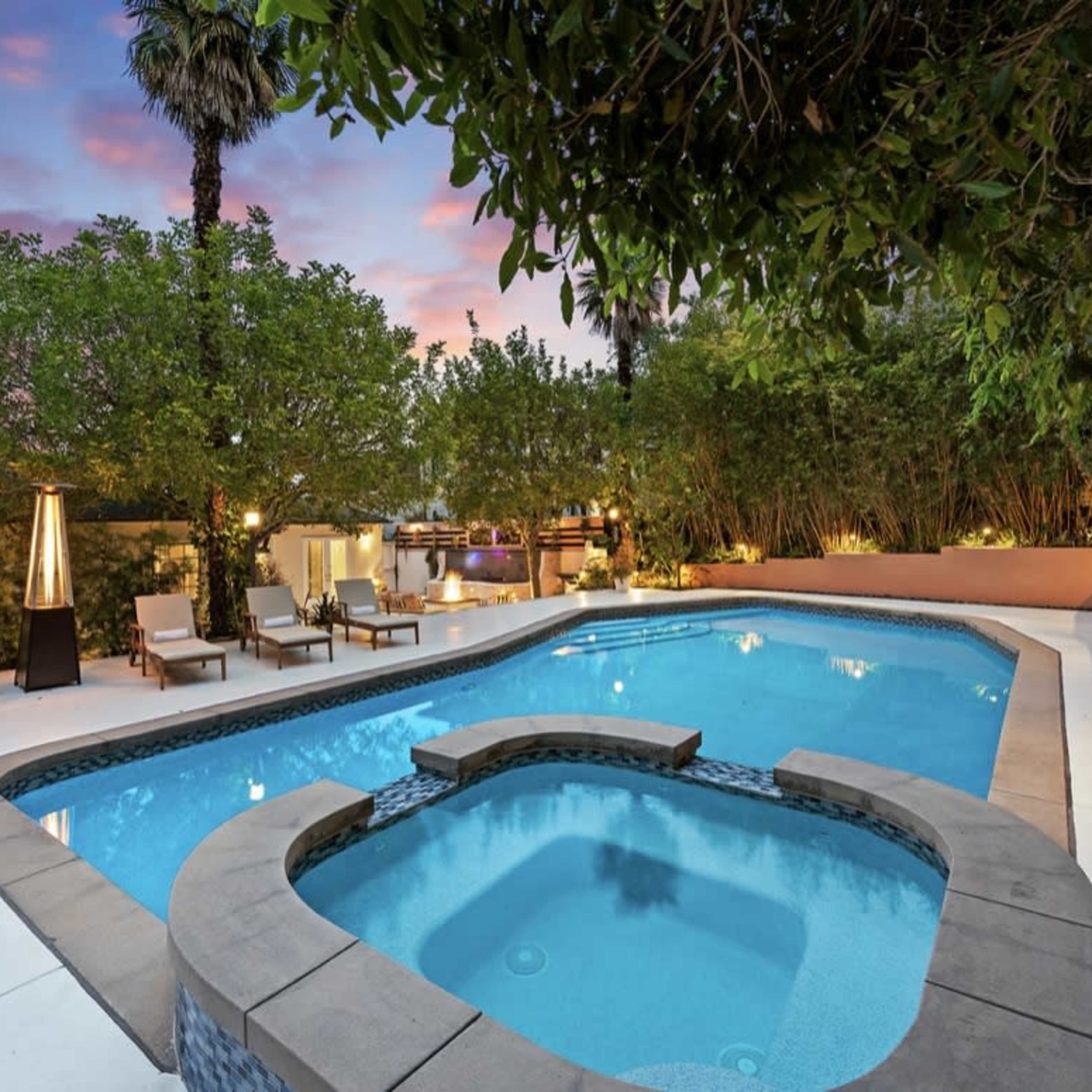 A tranquil backyard features a rectangular swimming pool with a spa, surrounded by lounge chairs and lush greenery under a sunset sky.