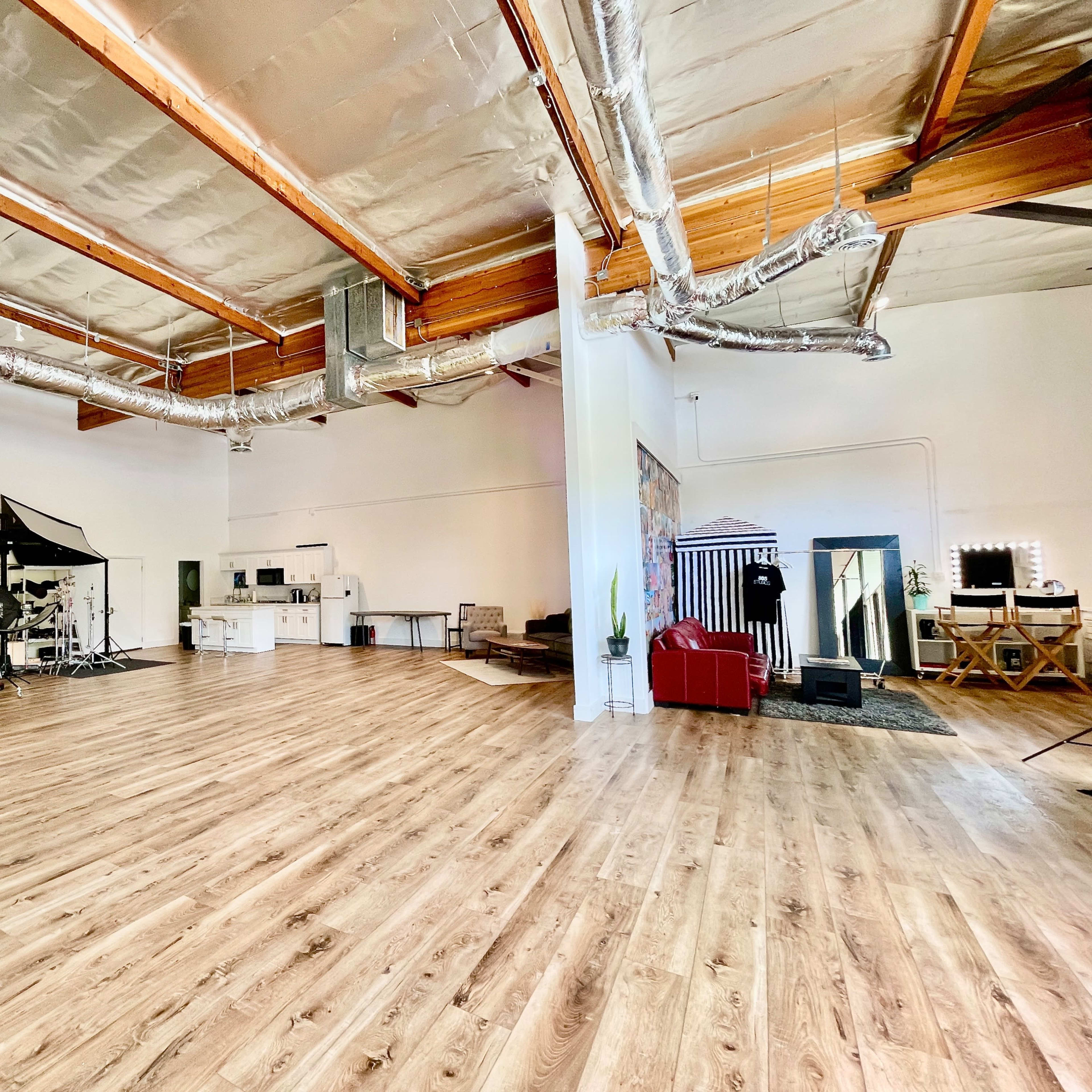 An expansive, well-lit studio space with wooden flooring, industrial-style ceiling beams, and various photography equipment positioned along one side.