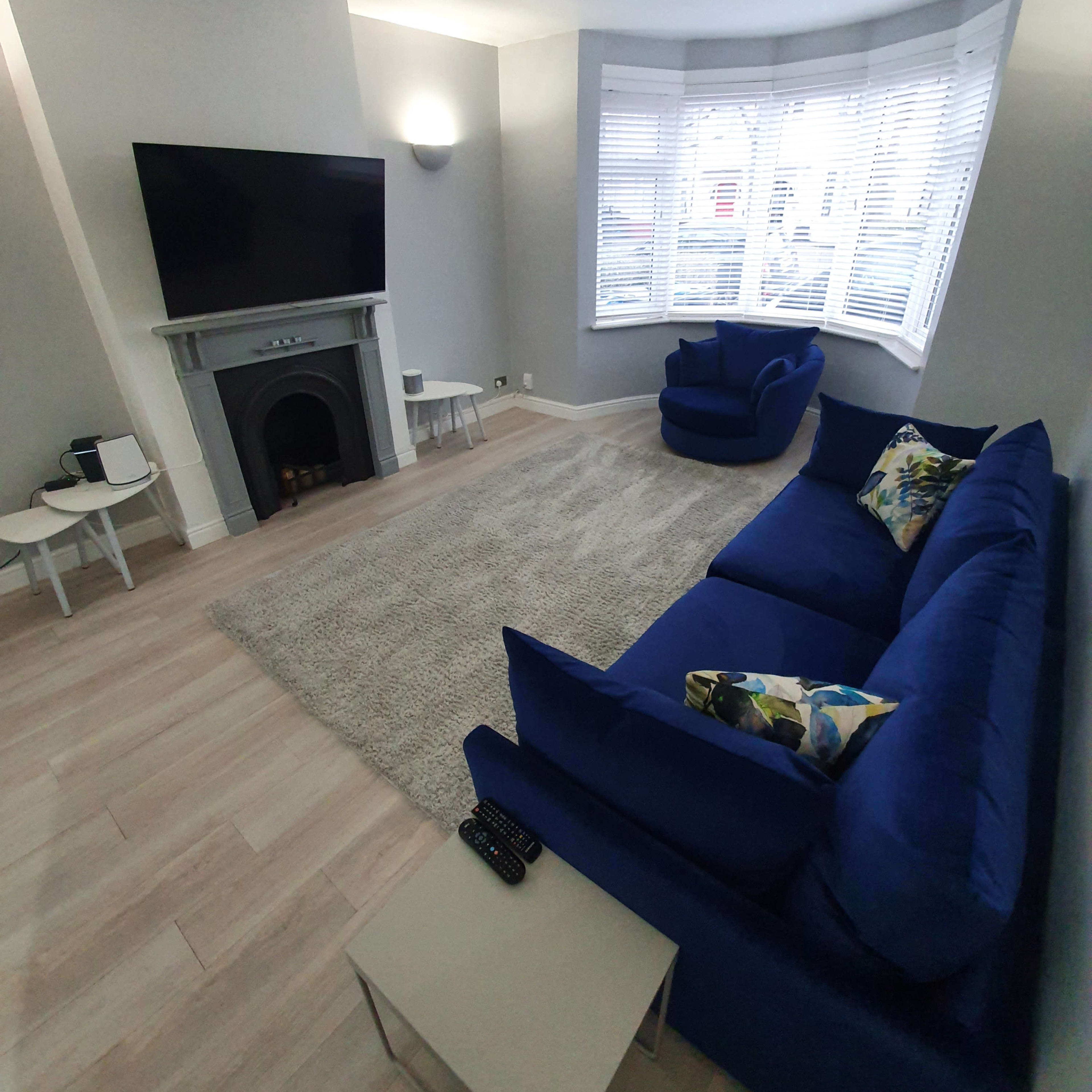 A modern living room featuring a blue sofa, two blue armchairs, a fireplace, and a large television mounted on a wall with ample natural light coming through bay windows.