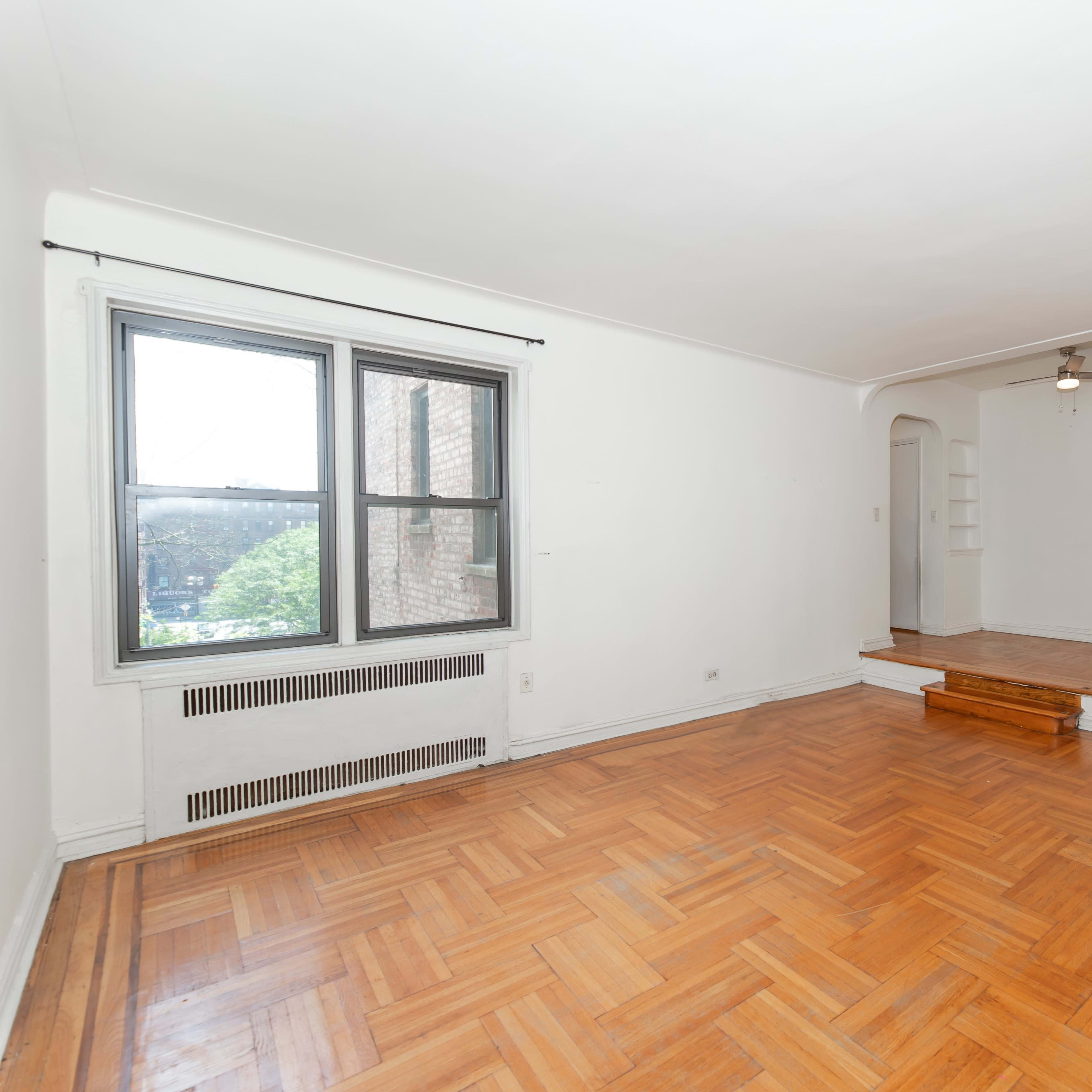 A vacant room with hardwood floors, a large window, and an entrance leading to a small alcove with a ceiling fan.
