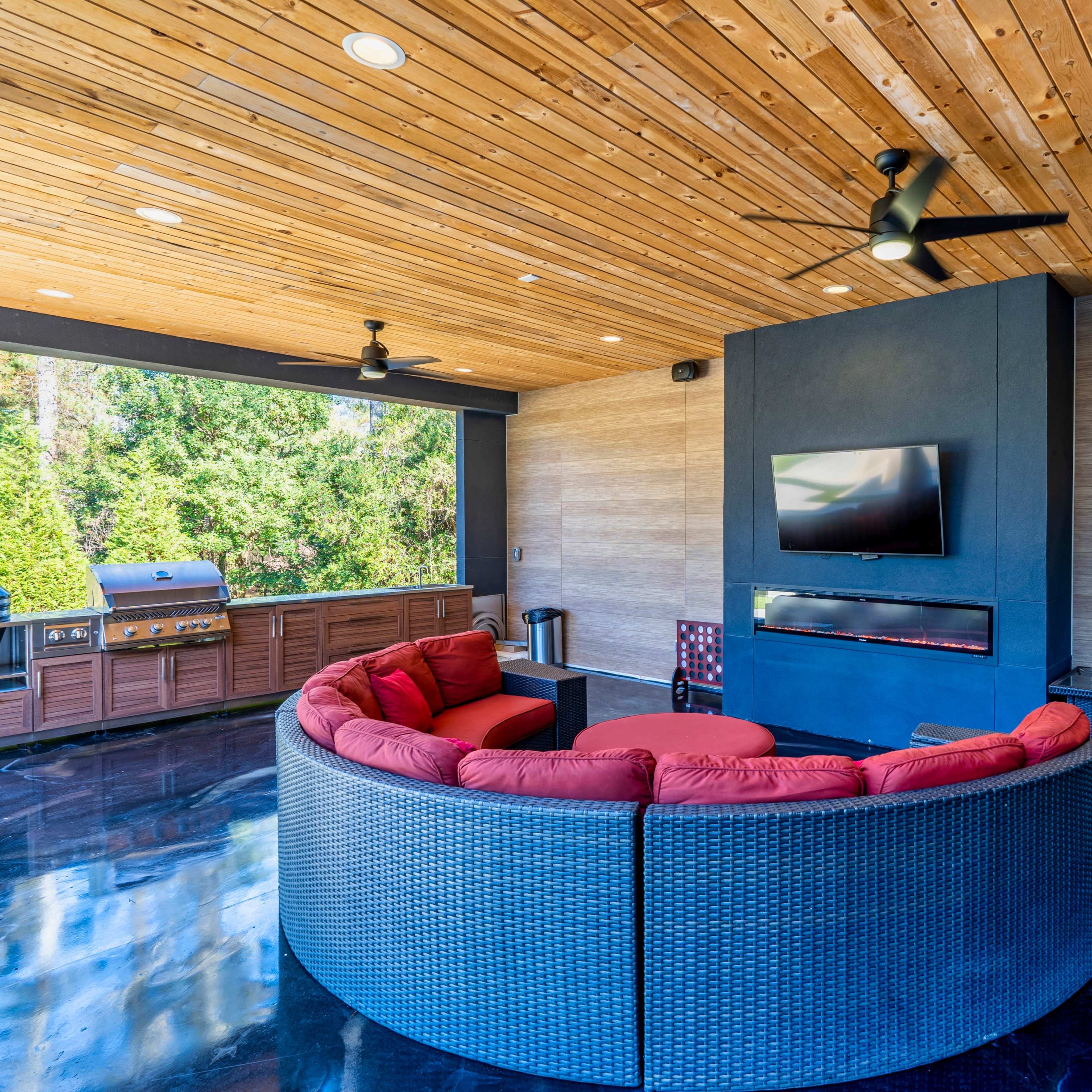 A modern outdoor living space featuring a curved red seating arrangement, a mounted television, a grill area, and large windows overlooking greenery.