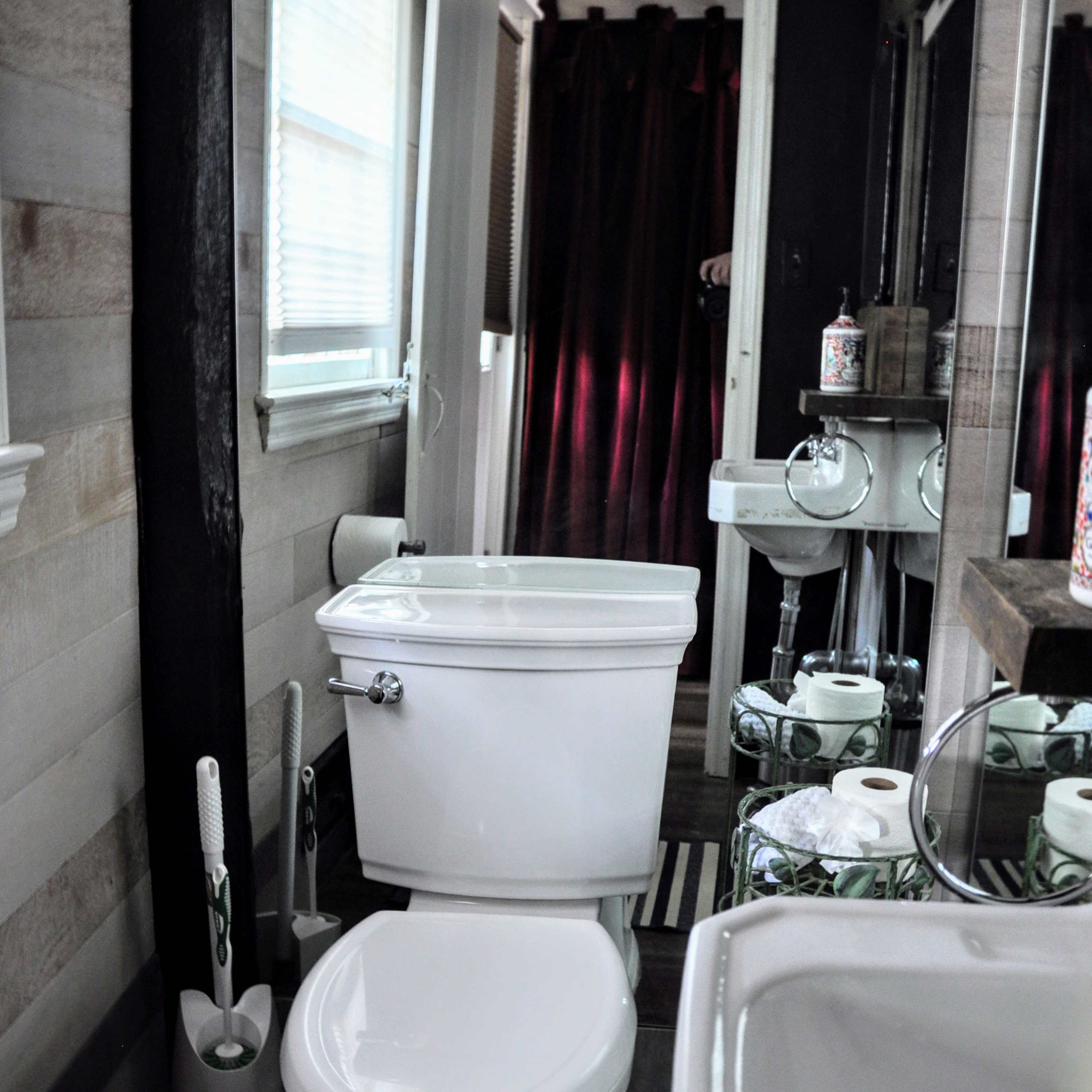 A narrow bathroom features a white toilet and sink along with a dark shower curtain, wooden wall accents, and a striped rug.