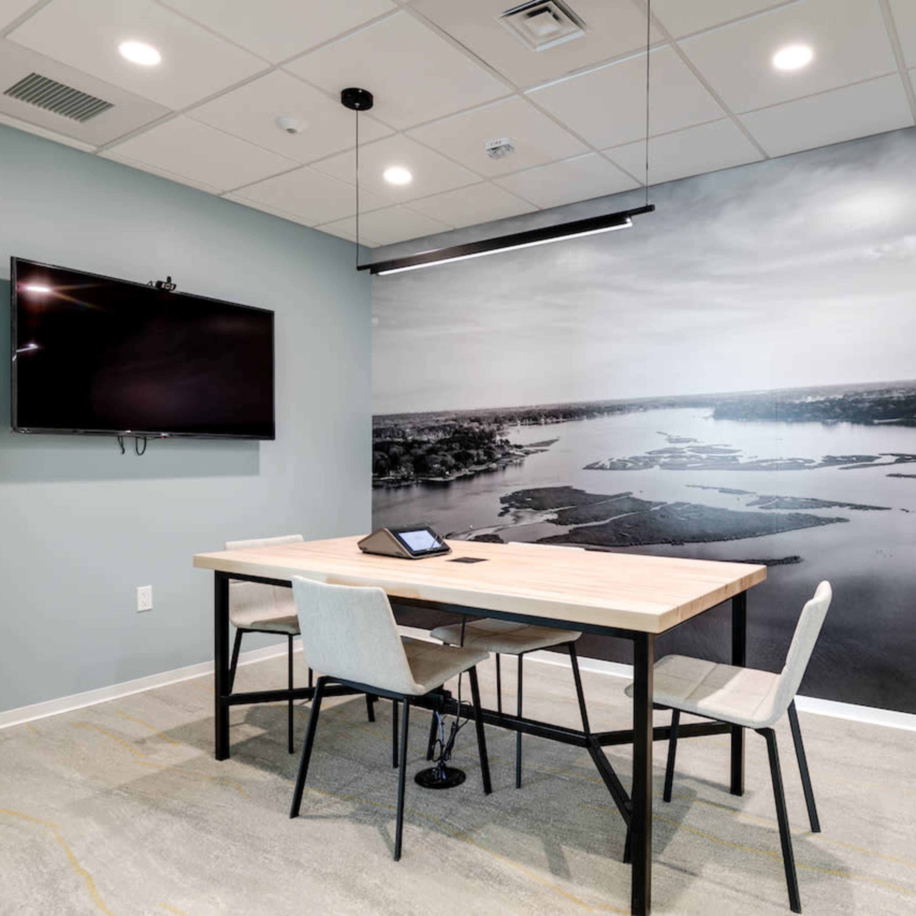 A modern conference room featuring a wooden table with four chairs, a mounted television, and a large black-and-white wall mural of a landscape.