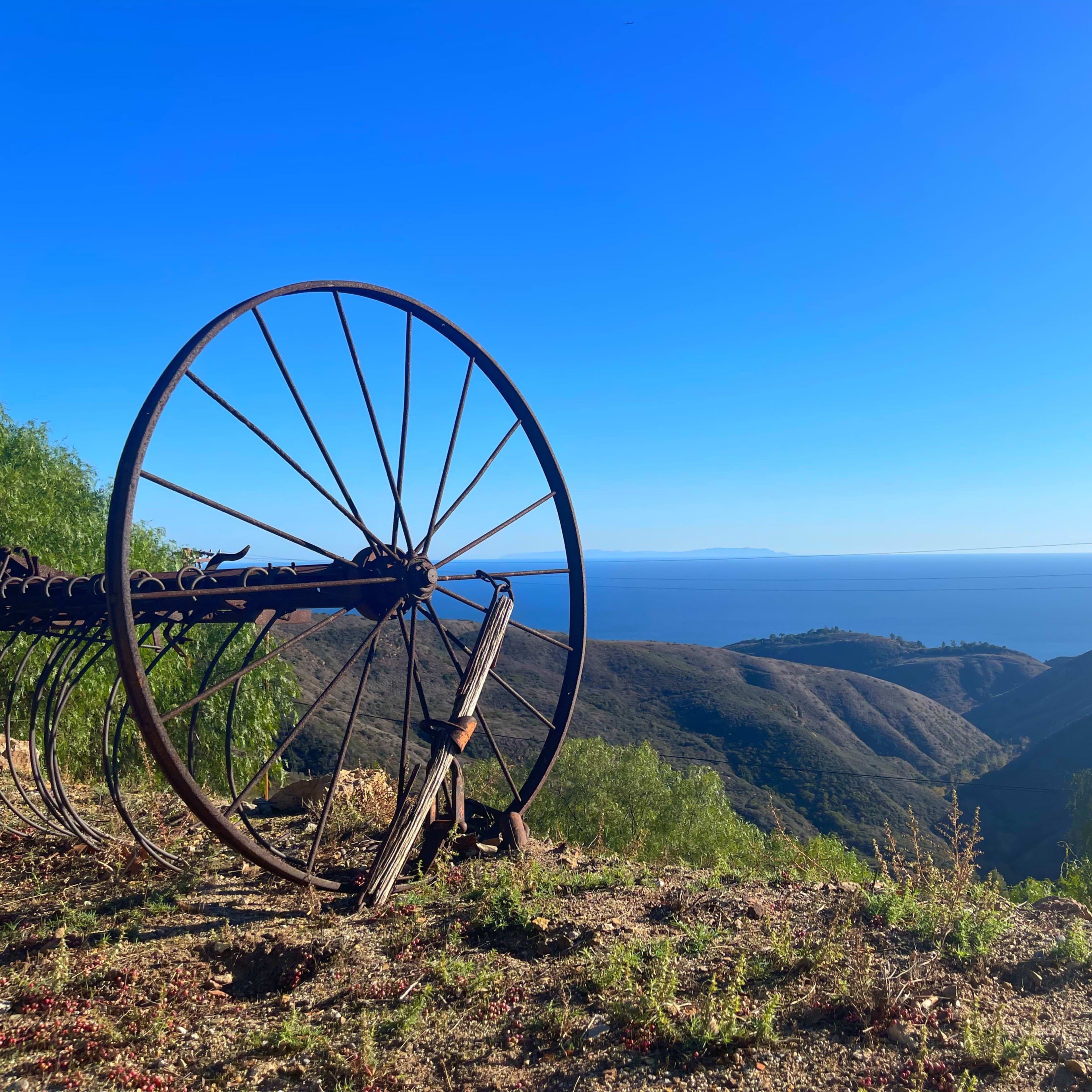 An old rusted wheel rests on a hillside overlooking the ocean and distant islands under a clear blue sky.
