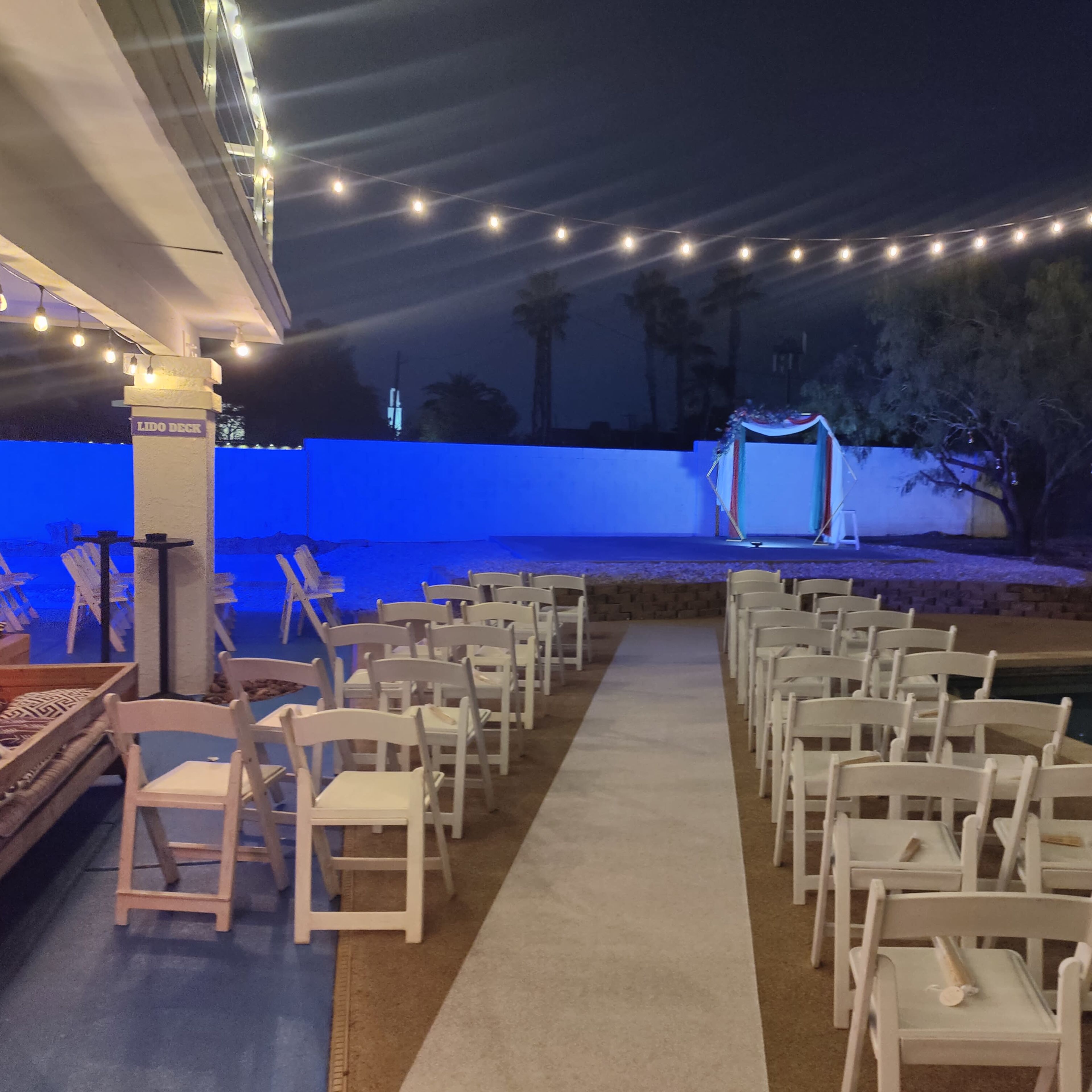 The scene shows a staged outdoor wedding ceremony with a row of white chairs lining a carpeted aisle under string lights, leading to a decorated archway.