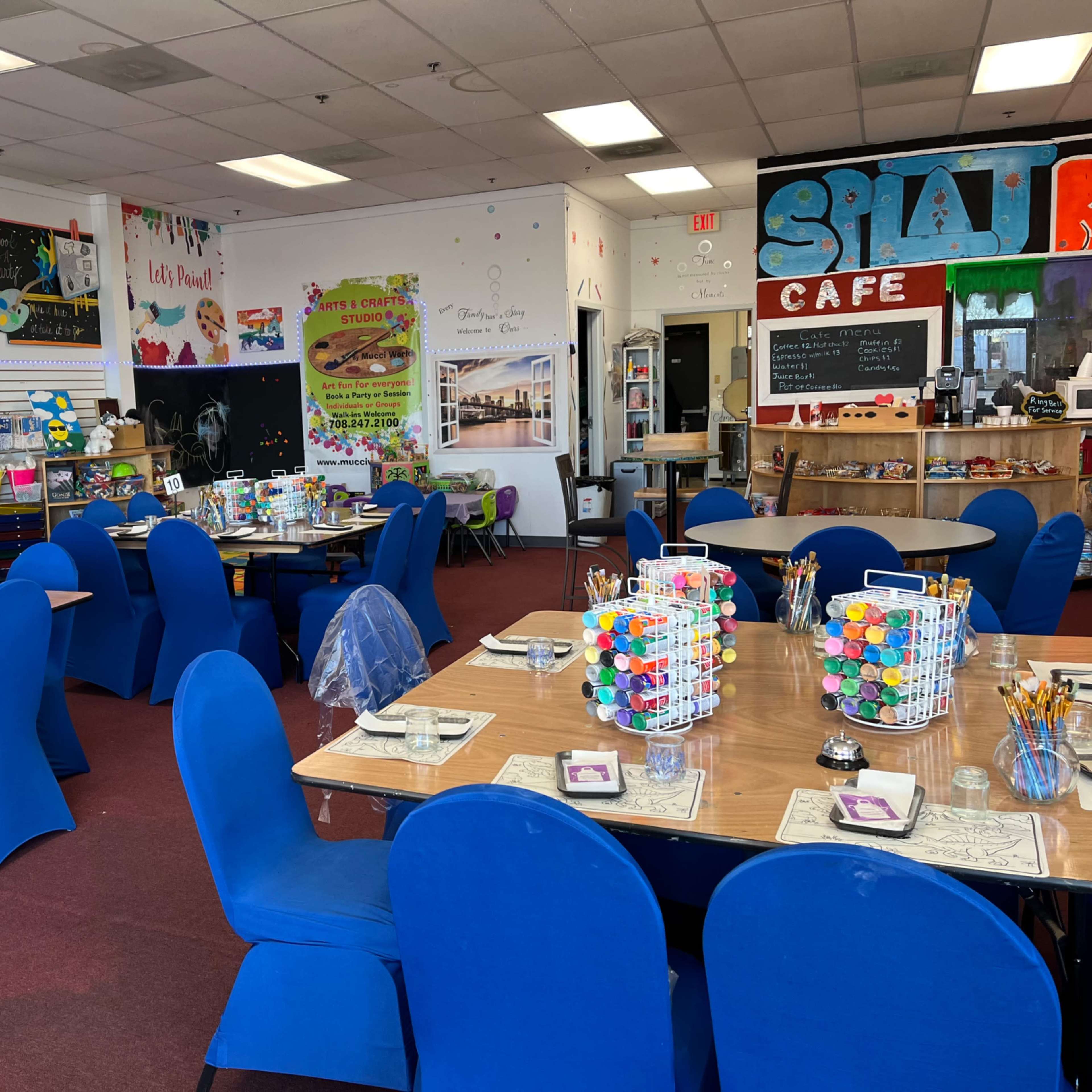 A brightly decorated art studio with blue chairs arranged around tables, each set with art supplies and colorful materials.