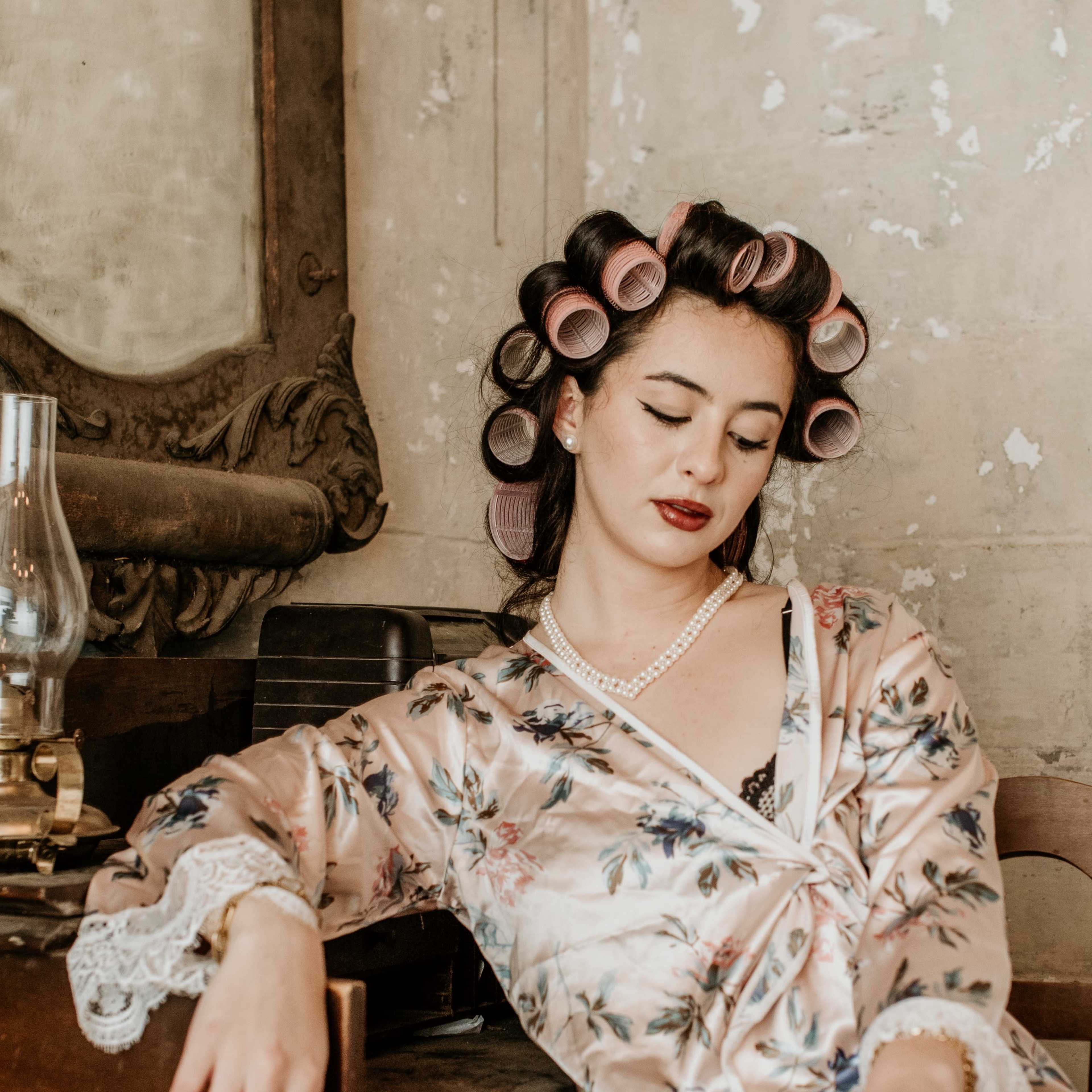 A woman in a floral robe sits at a vintage vanity with her hair in rollers, adorned with jewelry and red nail polish.