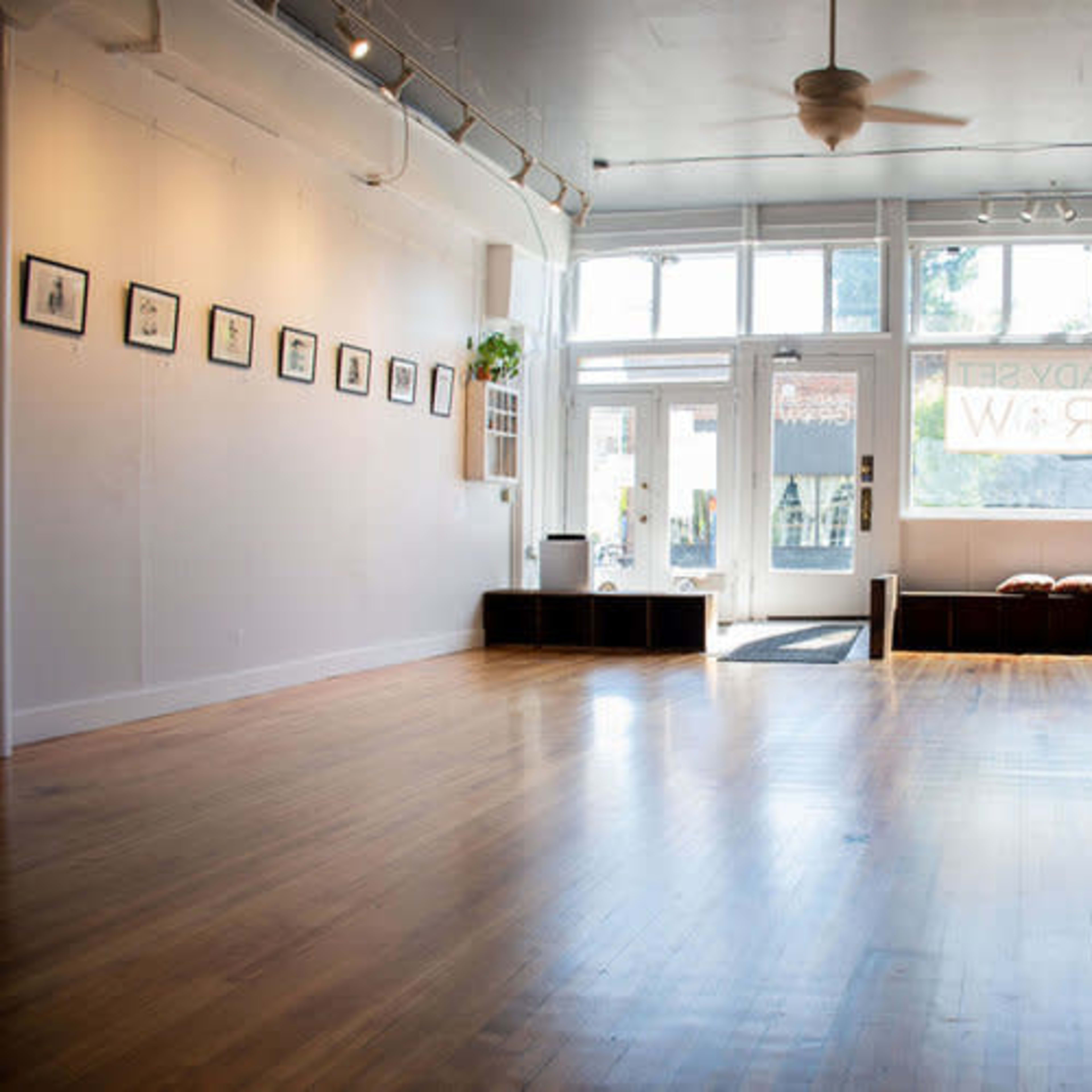 A spacious, brightly lit room with wooden flooring, featuring a few art frames on the wall and large windows letting in natural light.