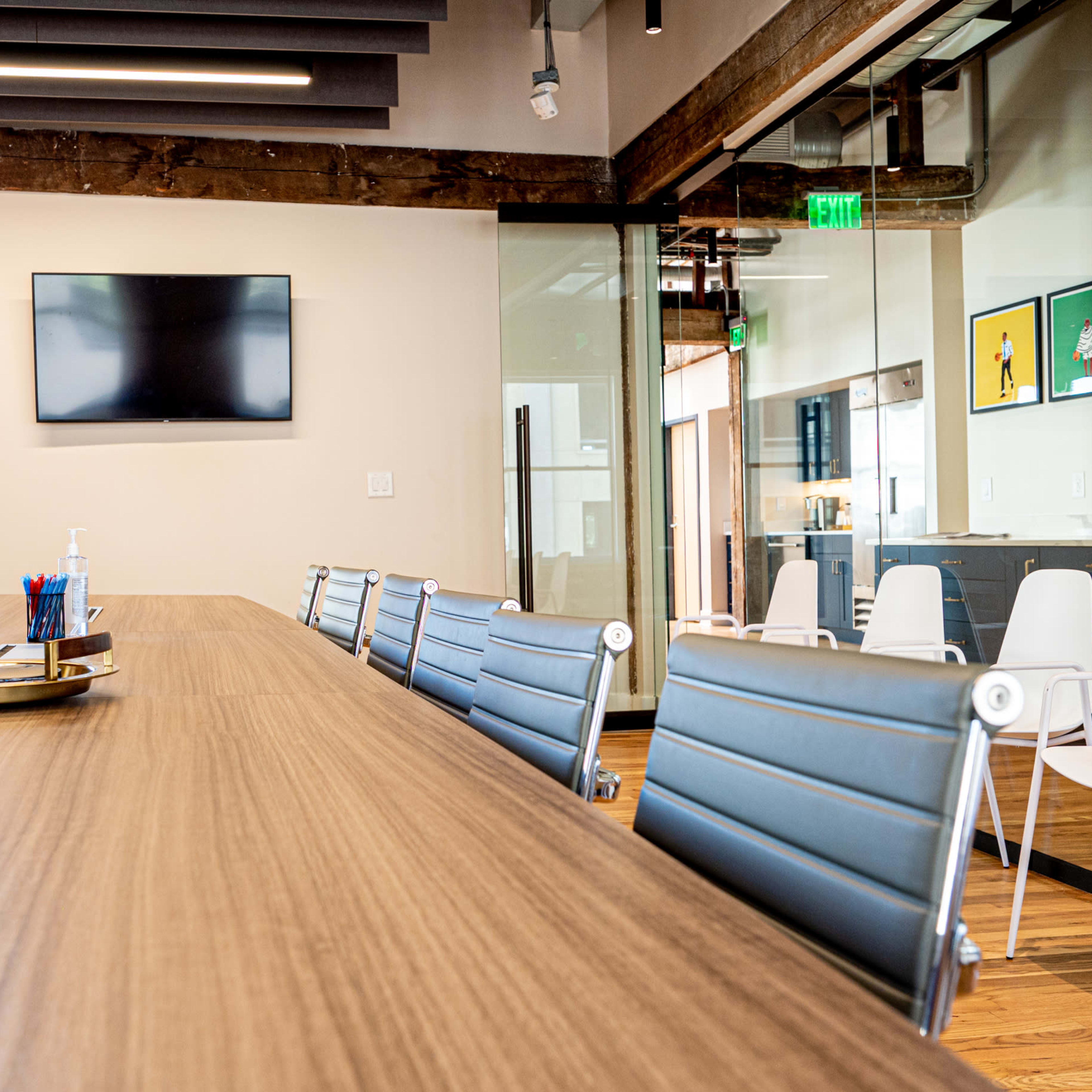 A modern conference room features a long wooden table, ergonomic chairs, a wall-mounted television, and large windows with exposed brick.