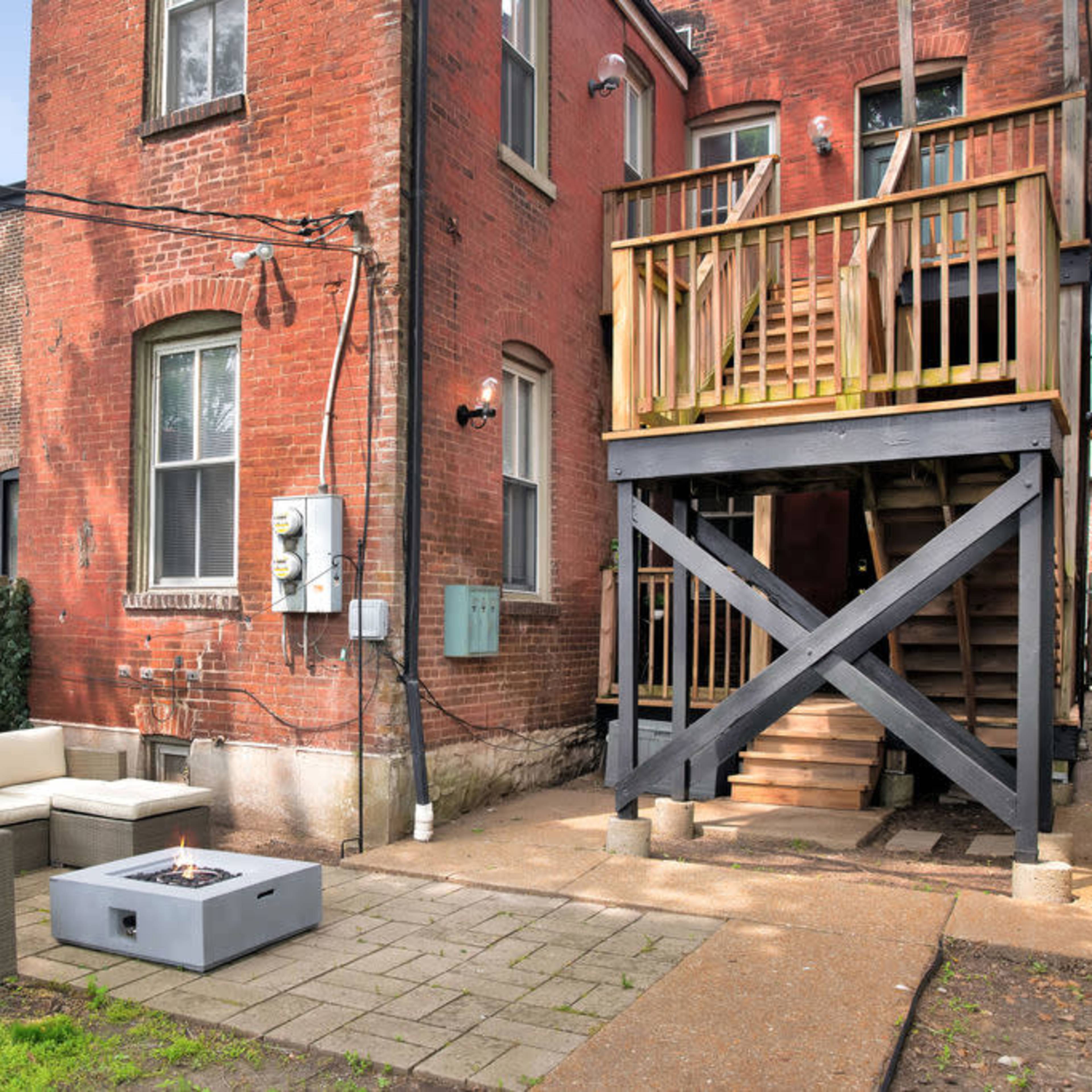 A backyard area with a seating arrangement and a fire pit near a brick building with a wooden staircase leading to an upper level.