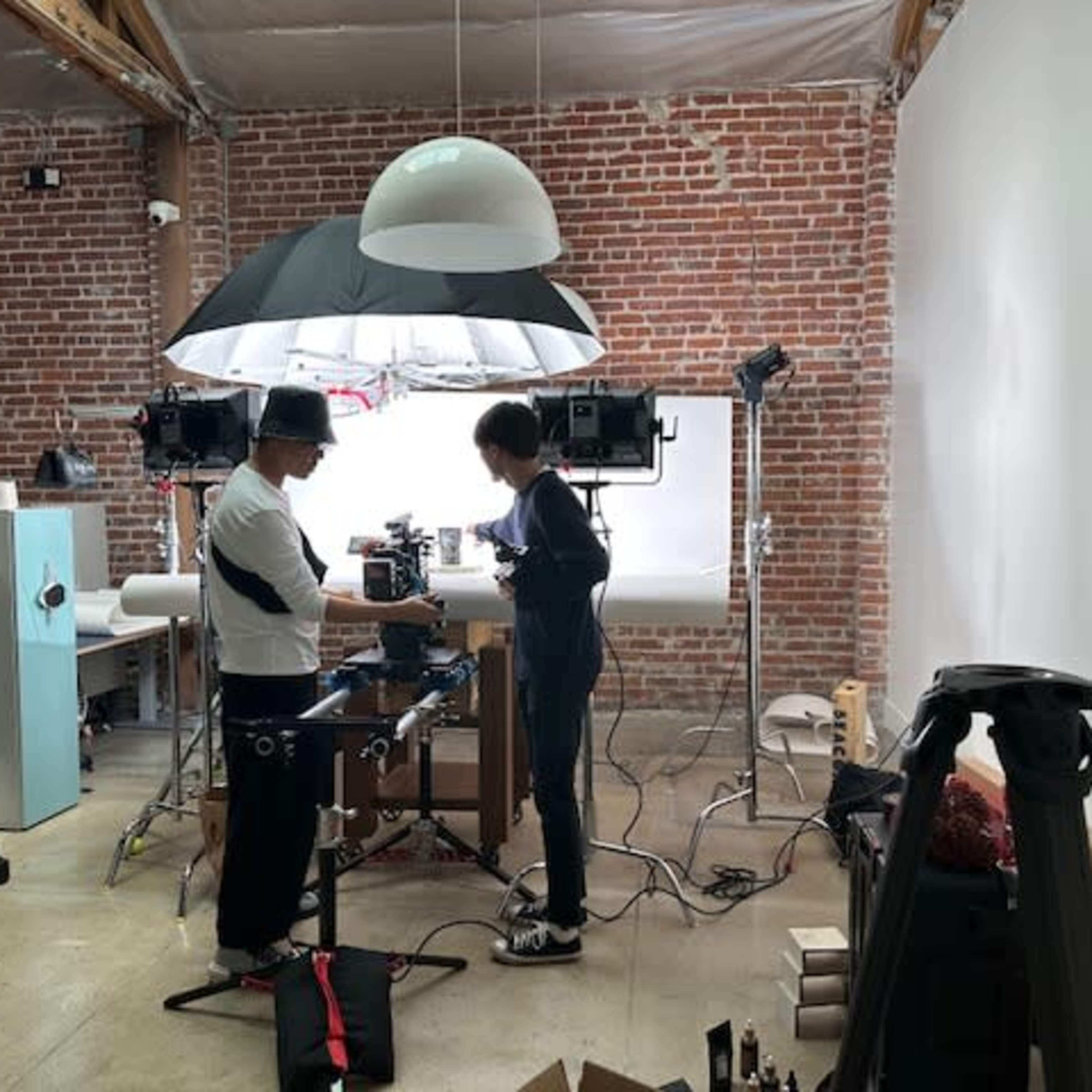 Two individuals are setting up a photography studio in a rustic brick-walled space, with lighting equipment and a backdrop in place.