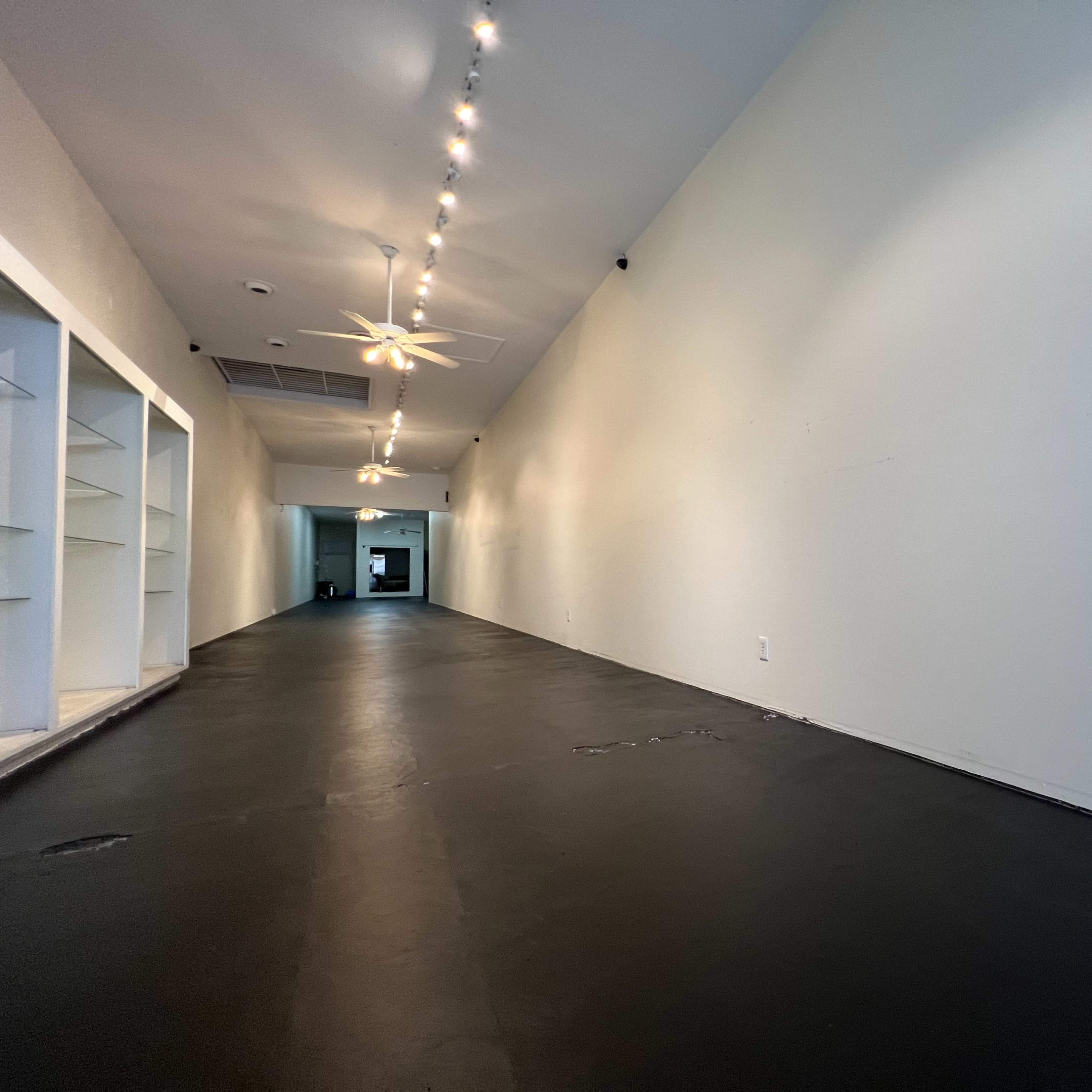 A long, narrow interior space with a black floor and white walls, illuminated by track lighting along the ceiling.