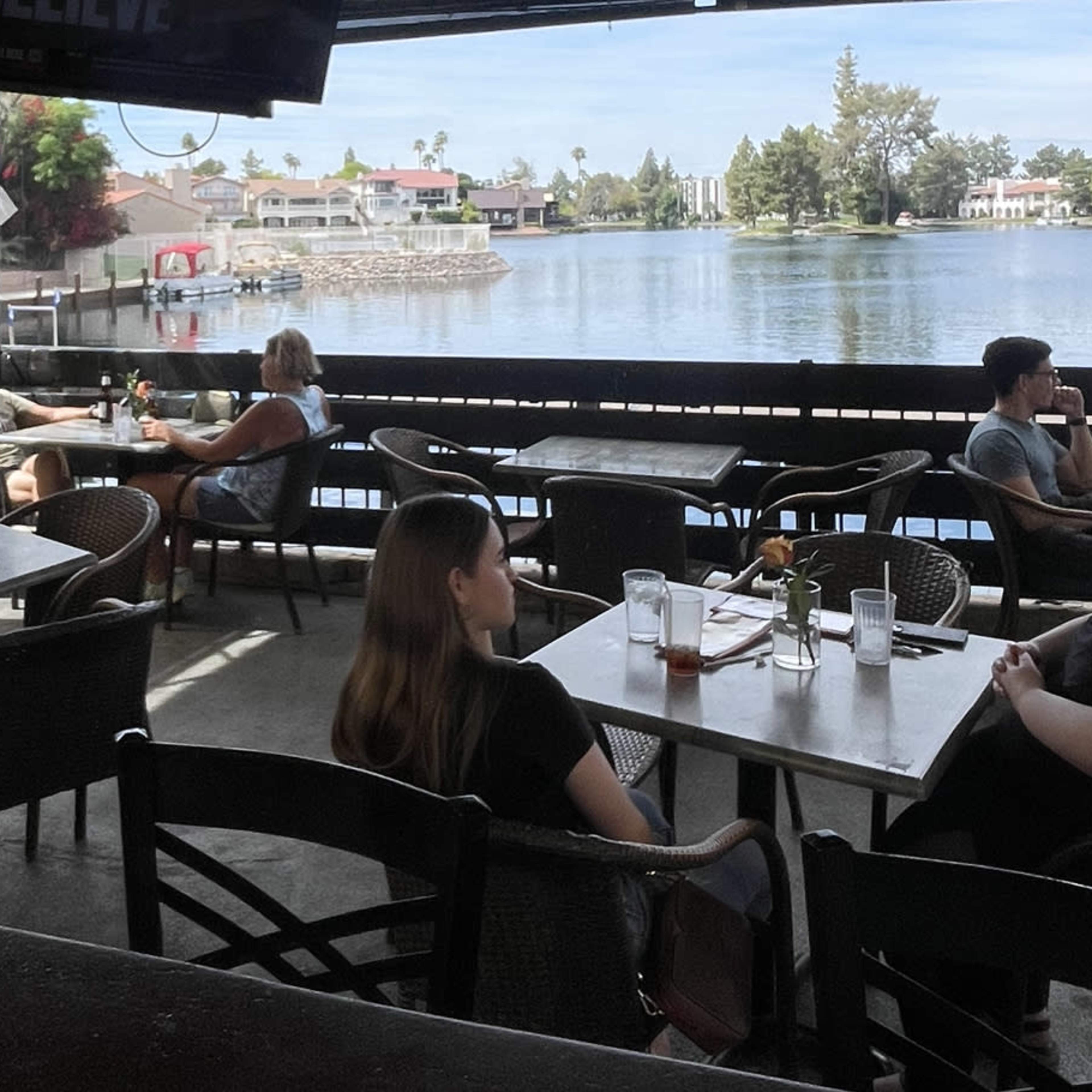 A lakeside restaurant patio with tables occupied by diners and a view of the water and homes in the background.