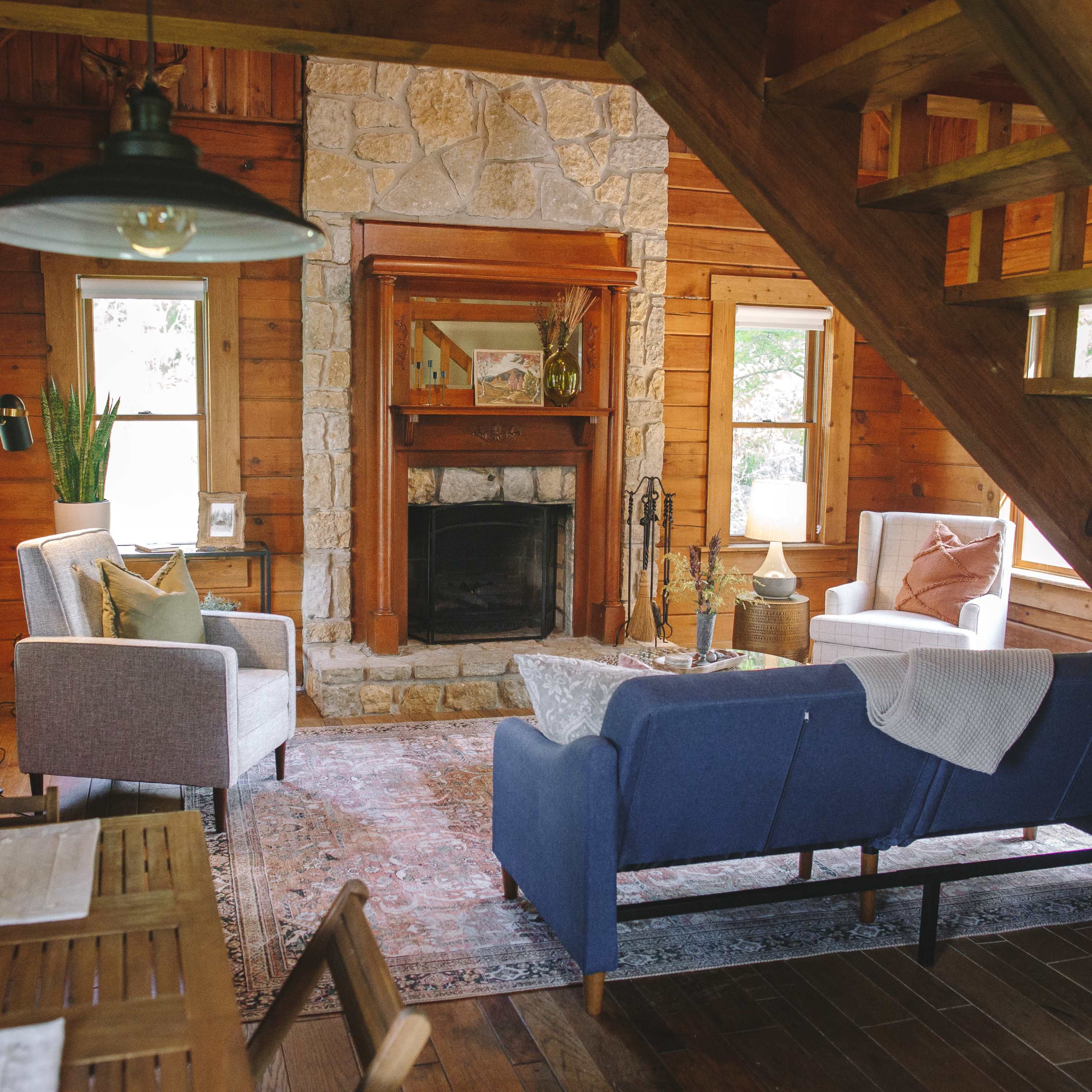 A cozy, wooden cabin interior featuring a living area with two chairs and a blue sofa arranged around a stone fireplace.