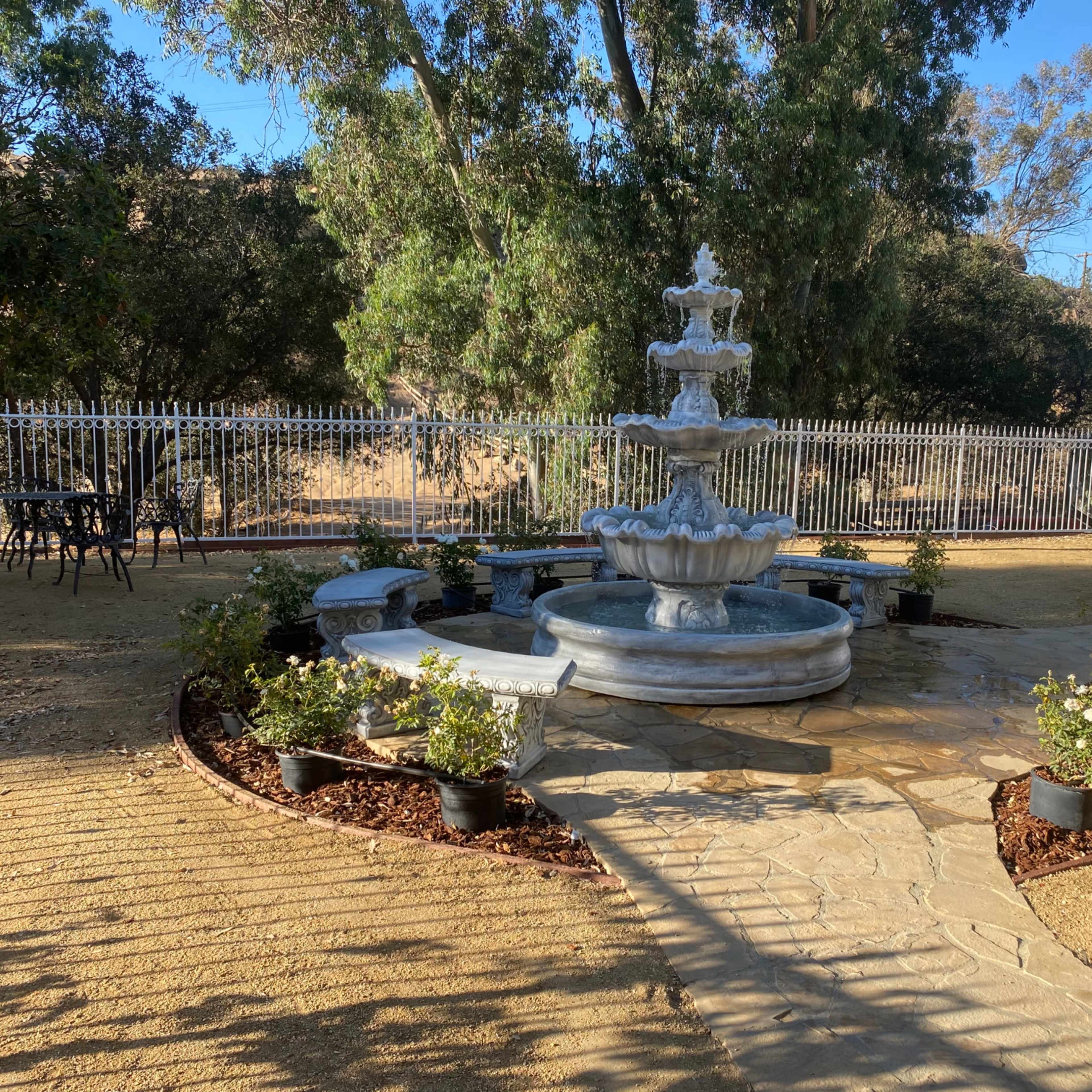 A three-tiered stone fountain is surrounded by potted plants and a stone pathway in a landscaped outdoor area.