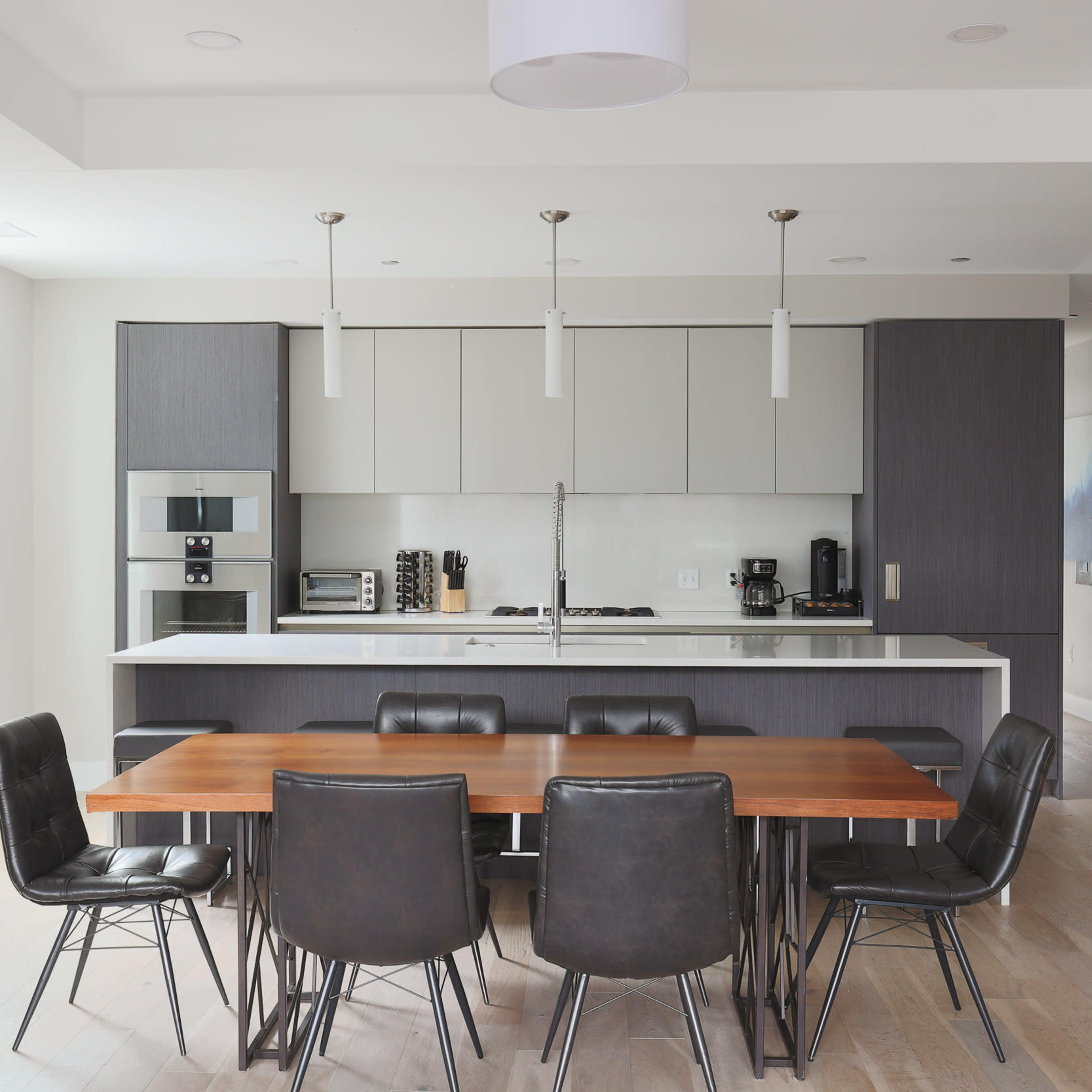 A modern kitchen and dining area features a long wooden table surrounded by black leather chairs, with a sleek kitchen cabinet and appliances in the background.