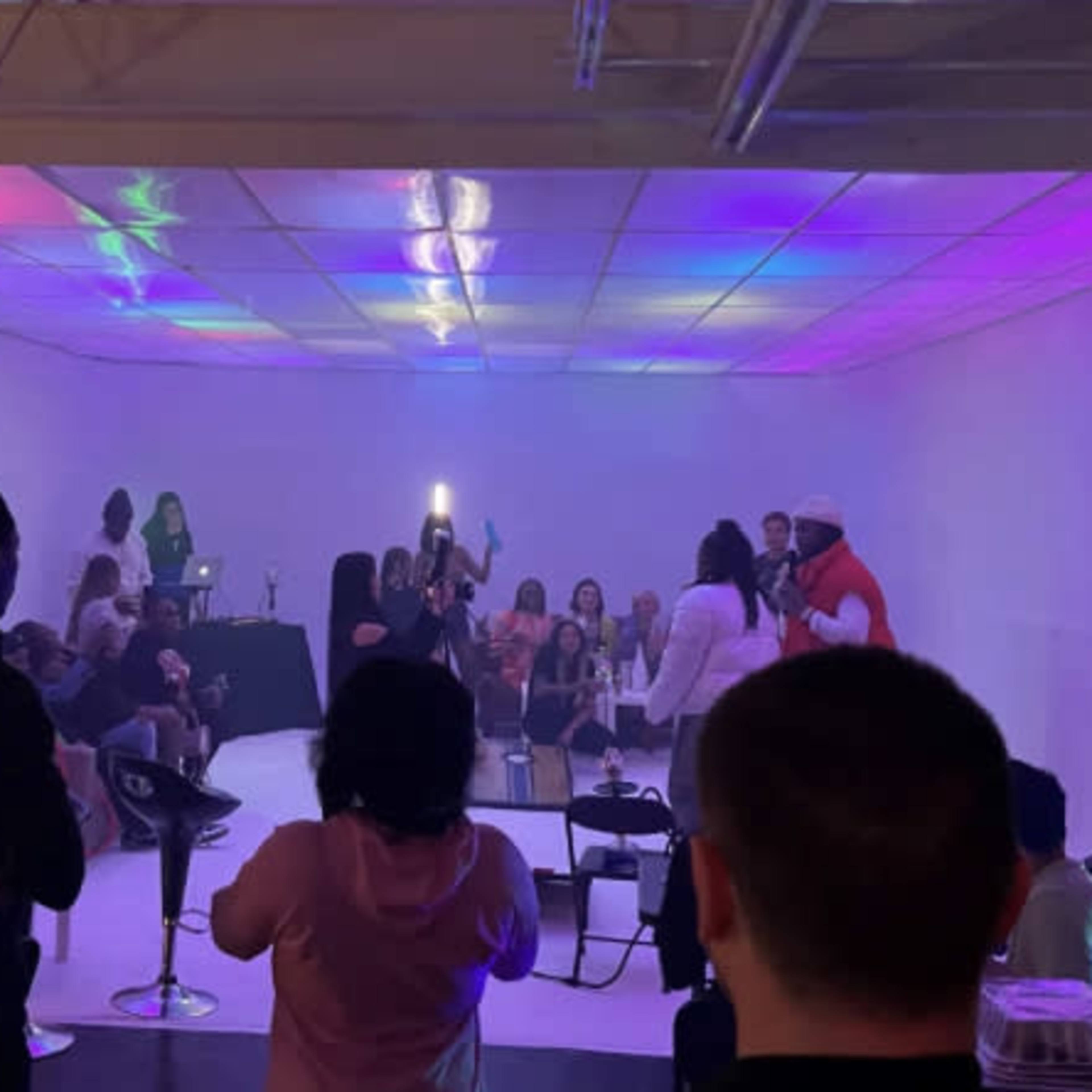 A group of people is gathered in a brightly lit room with colorful lights and chairs, while some individuals appear to be performing or presenting on a small stage.