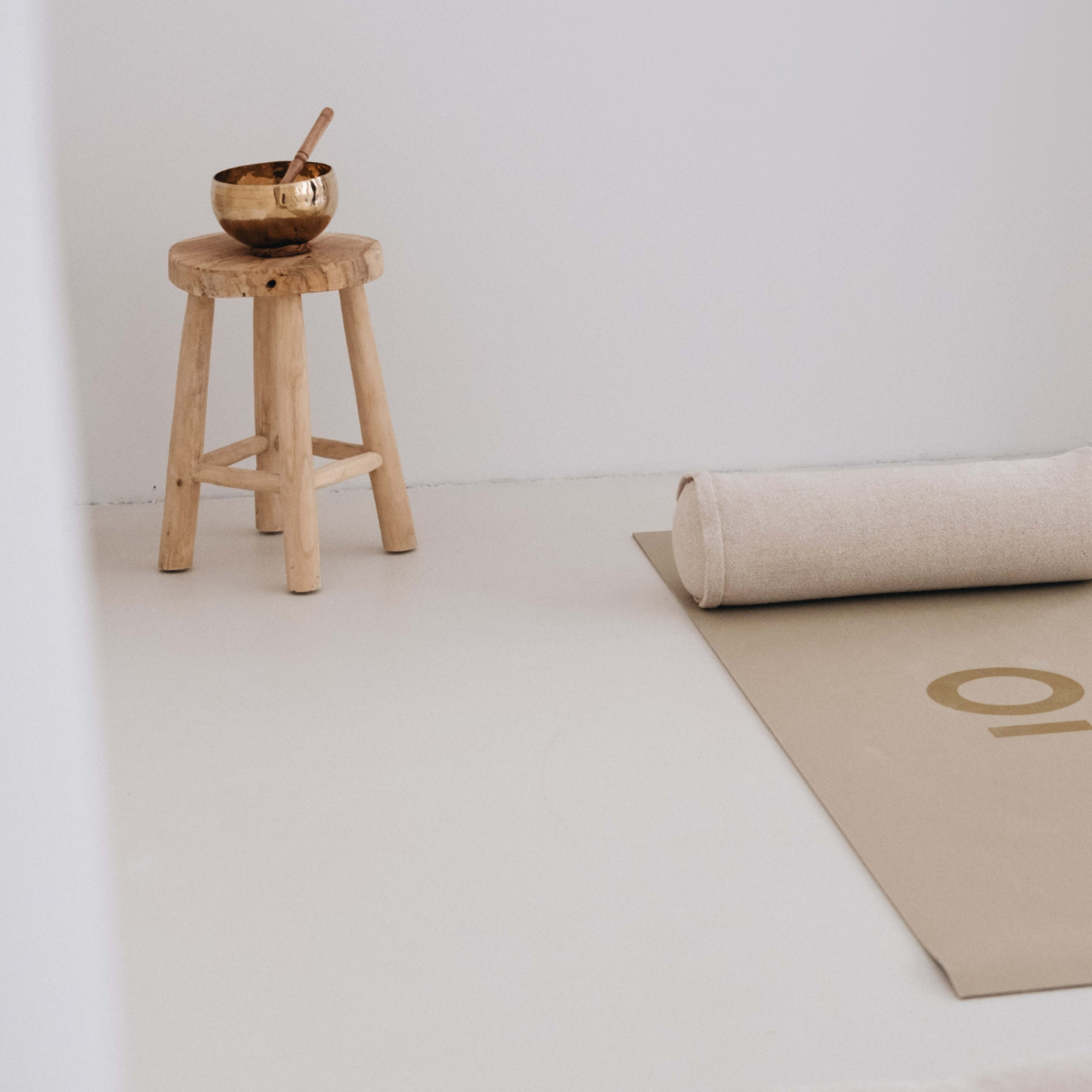 A rolled yoga mat with the number "10" is positioned next to a wooden stool and a metal bowl in a minimalistic room.