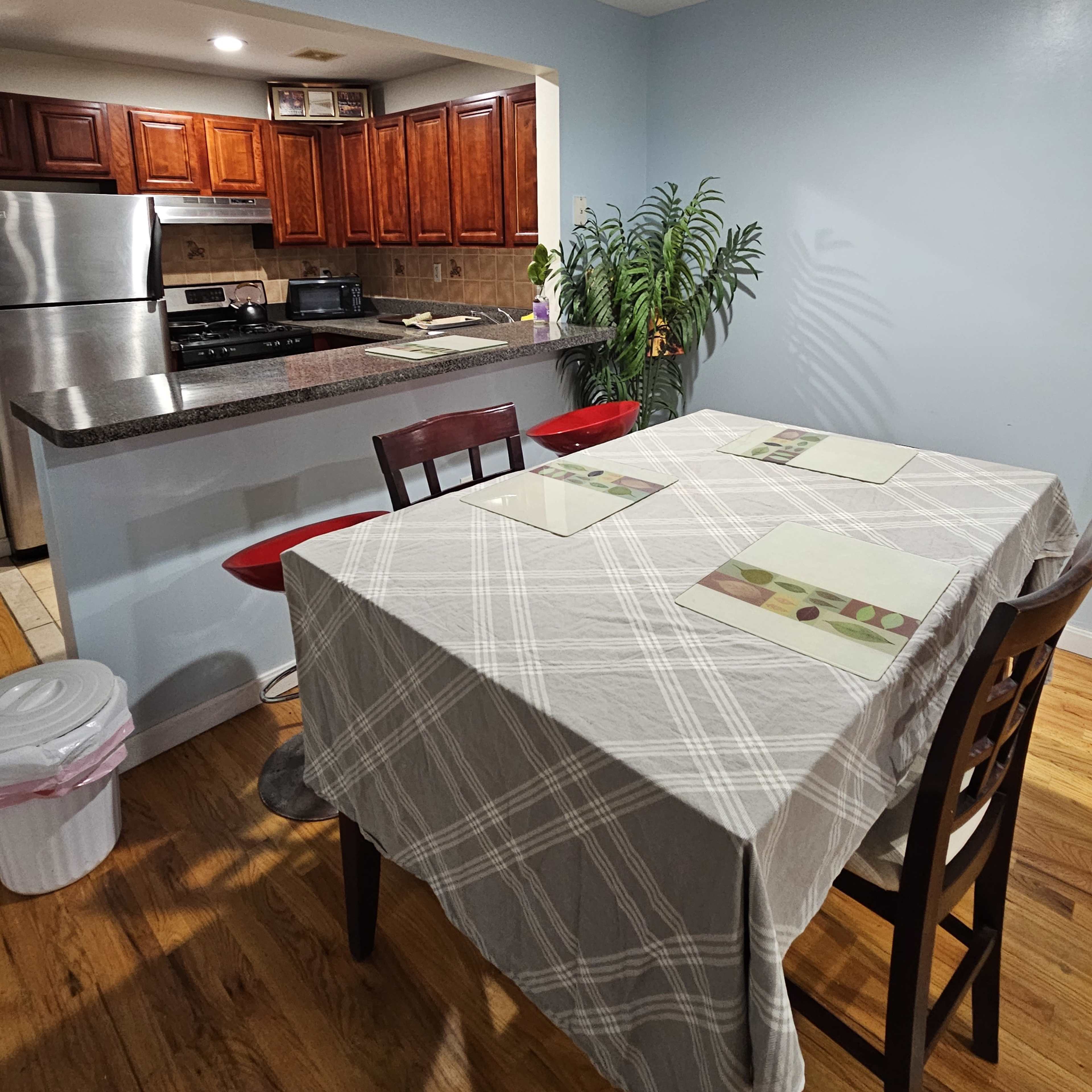 A dining table covered with a checkered tablecloth is set for a meal in a kitchen with wooden cabinets and stainless steel appliances.