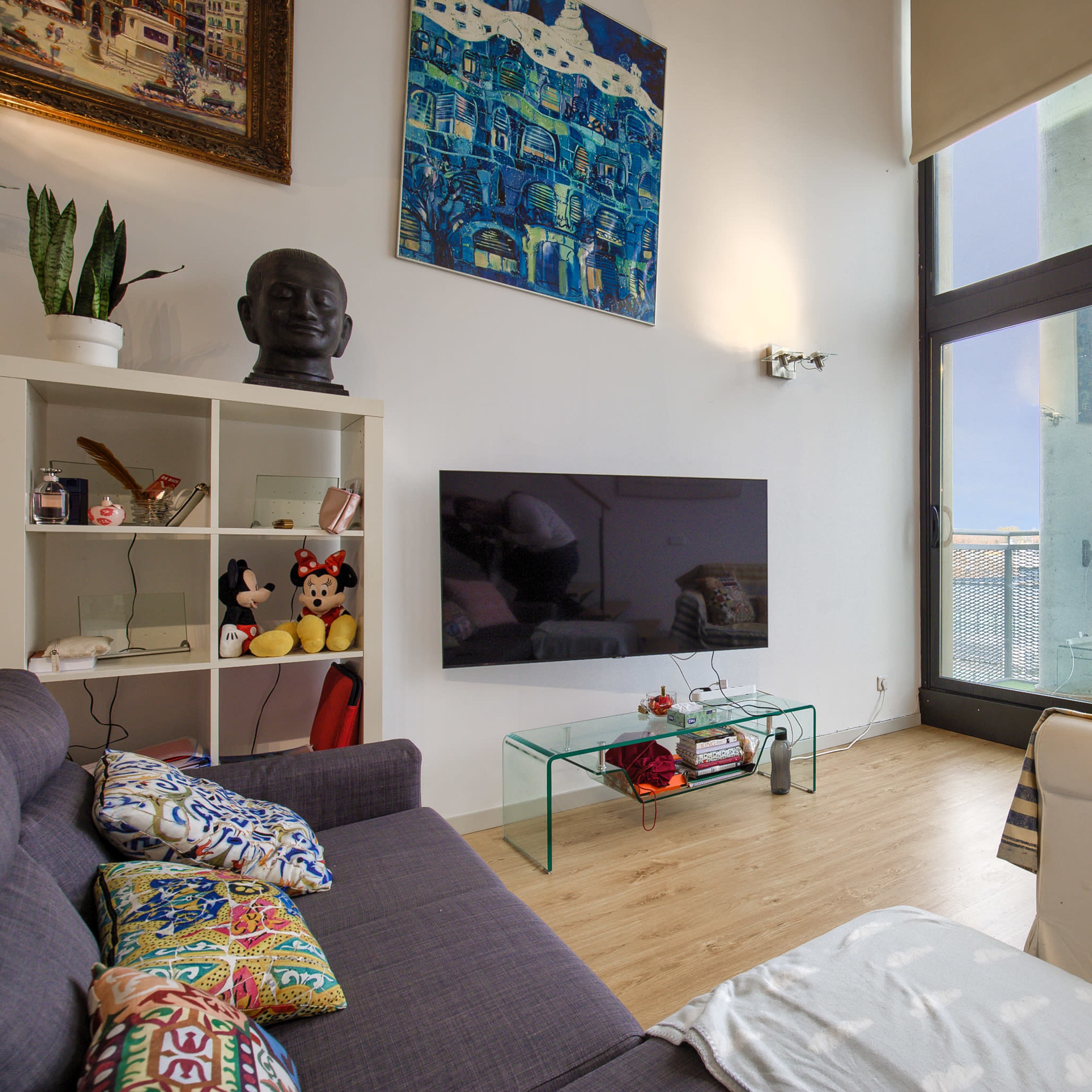 A modern living room with a large window overlooking a balcony, featuring a television on a glass stand, a bookshelf, and two sofas with decorative cushions.