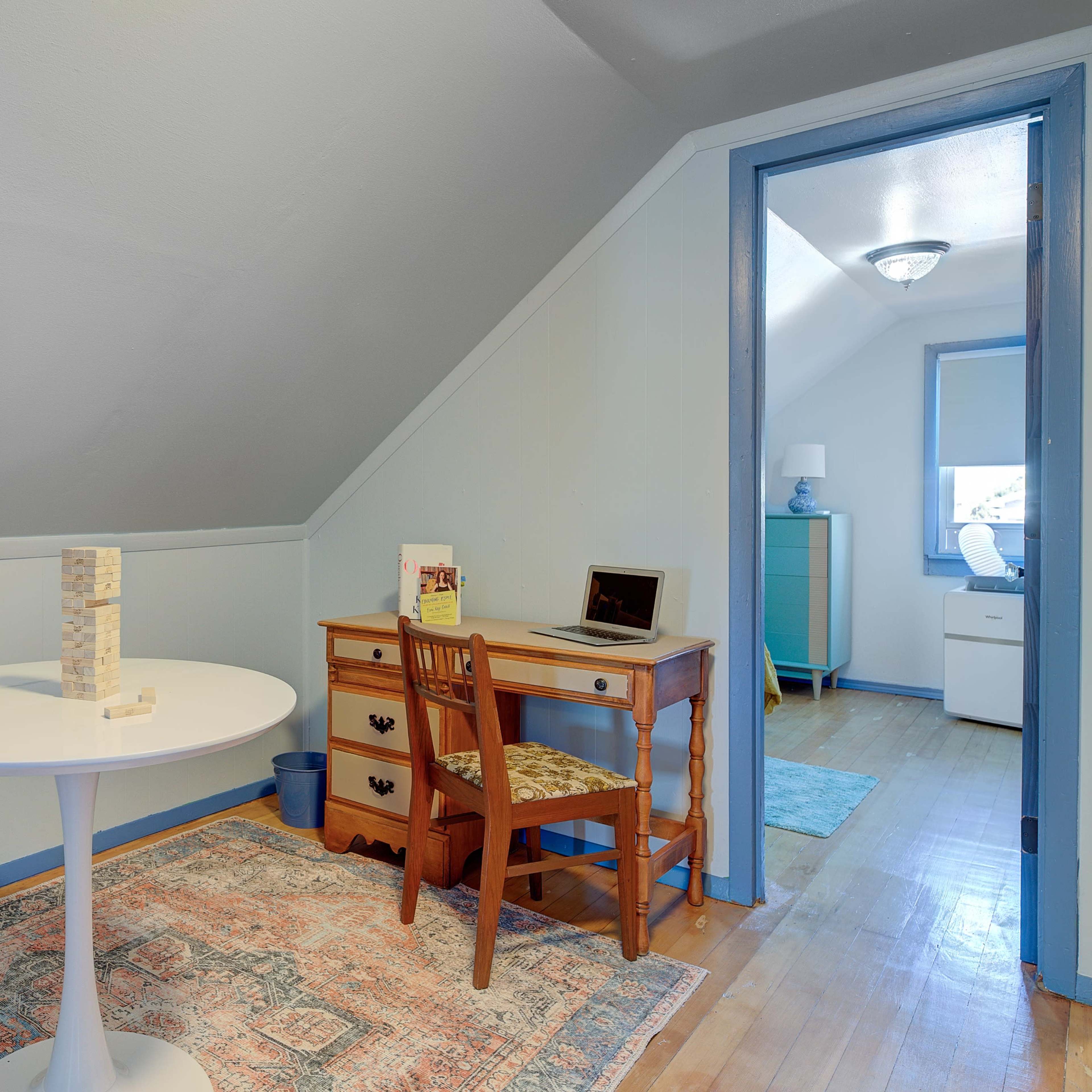 A small room with a sloped ceiling, featuring a round white table, a wooden desk with a chair, and a rug on the hardwood floor.