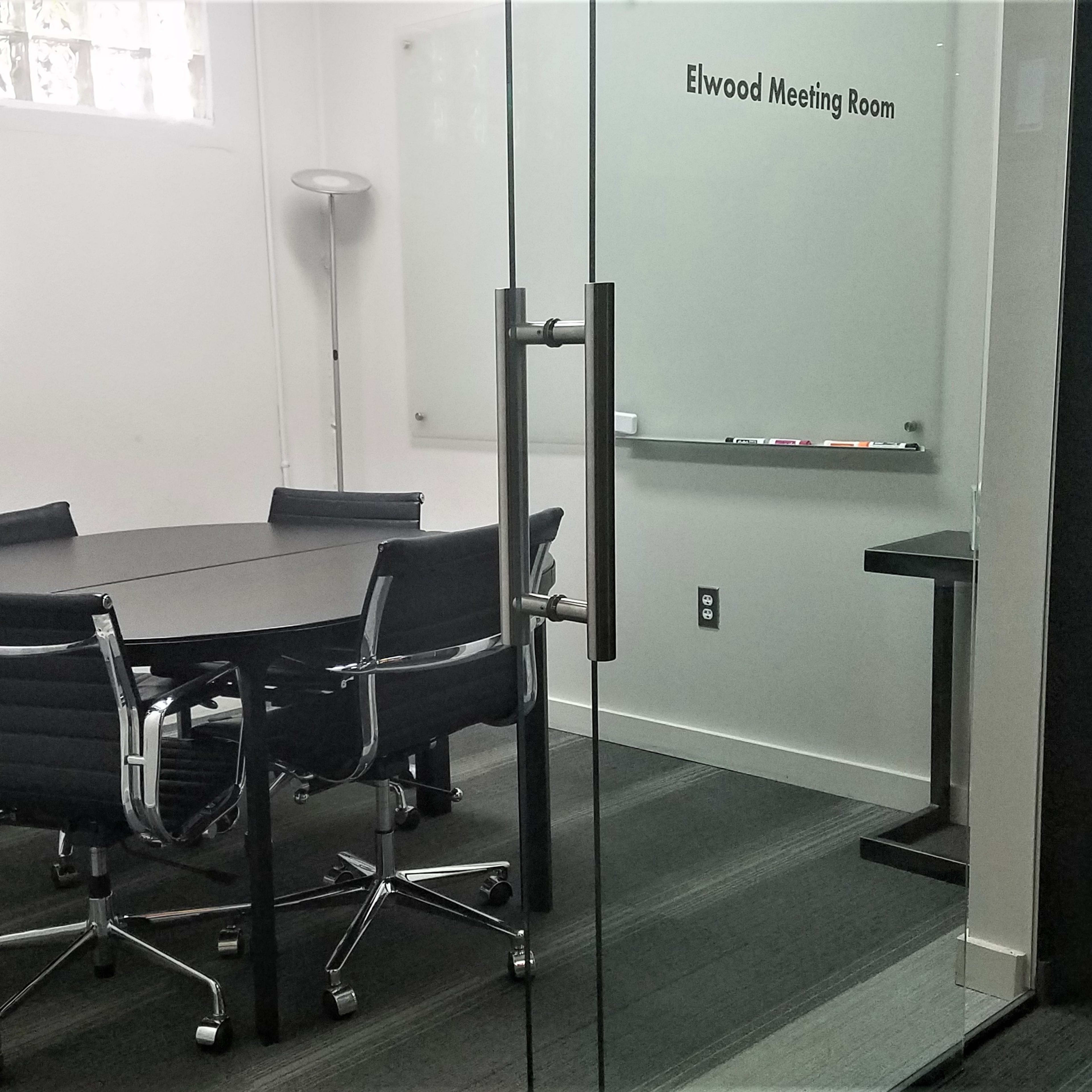 A modern meeting room with a round table, six chairs, a television on the wall, and a whiteboard.