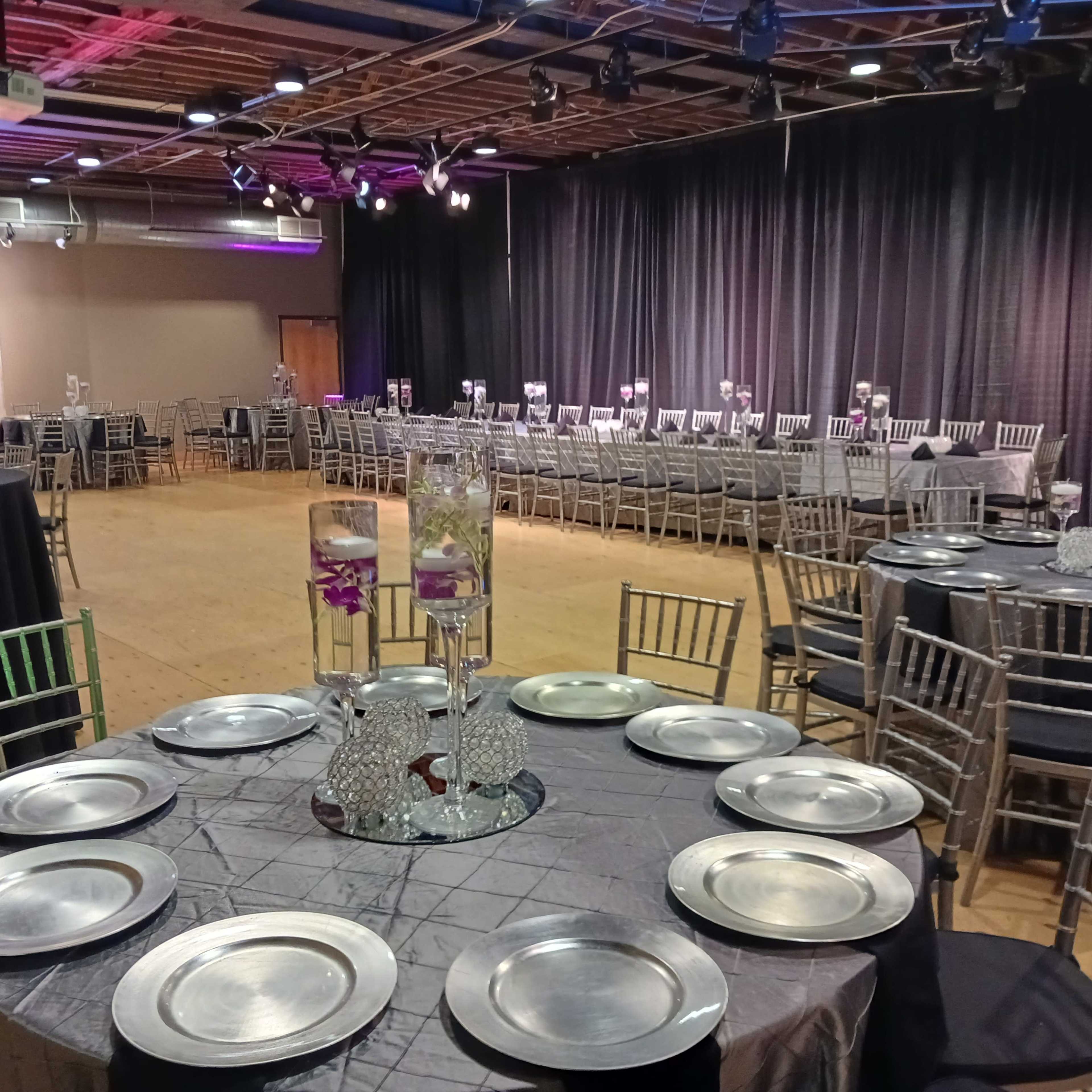 A large event space features elegantly set tables with silver plates and centerpieces, arranged for a formal gathering.