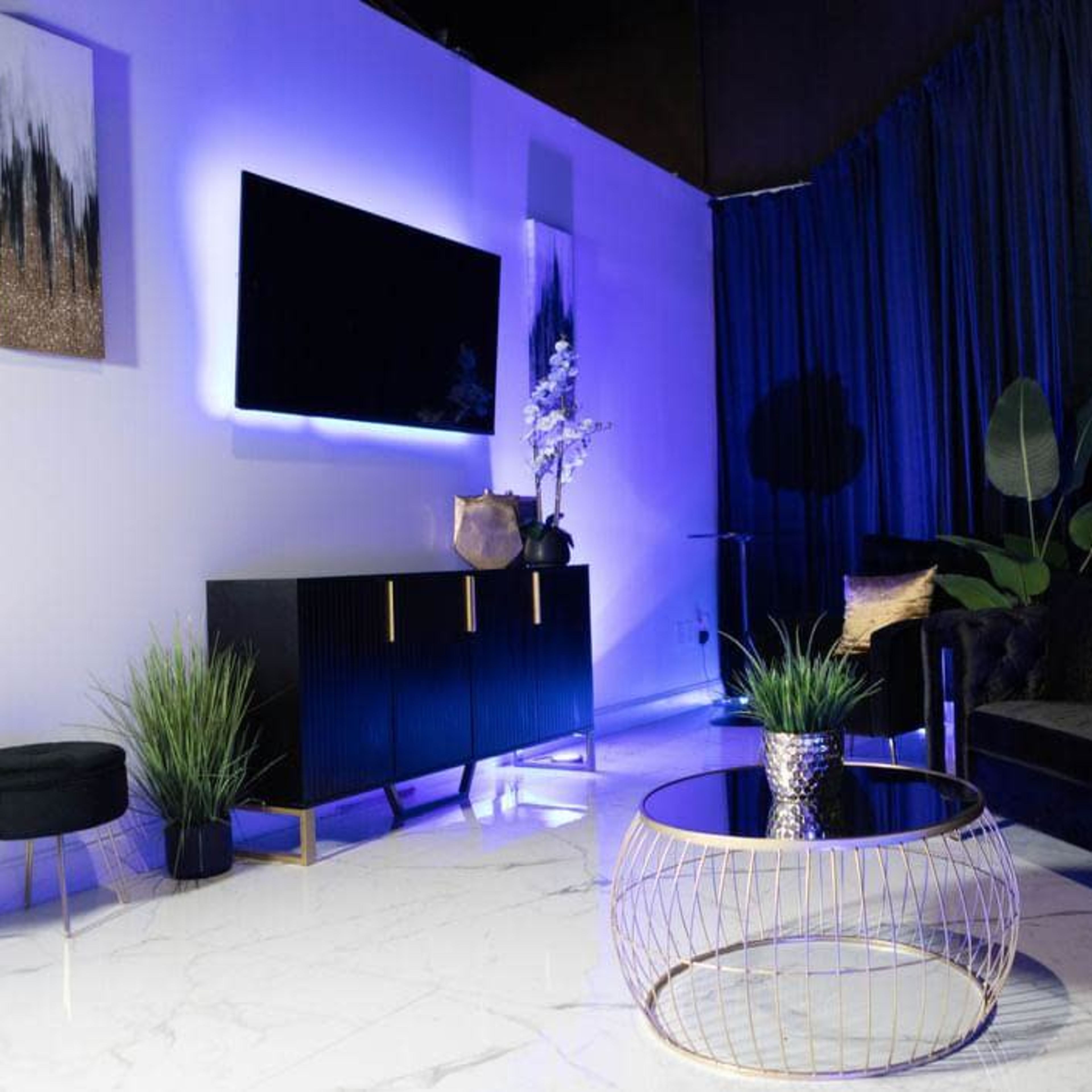 A modern living room with a black sofa, a round coffee table, a large television mounted on a white wall, and blue lighting accents against a backdrop of dark curtains.