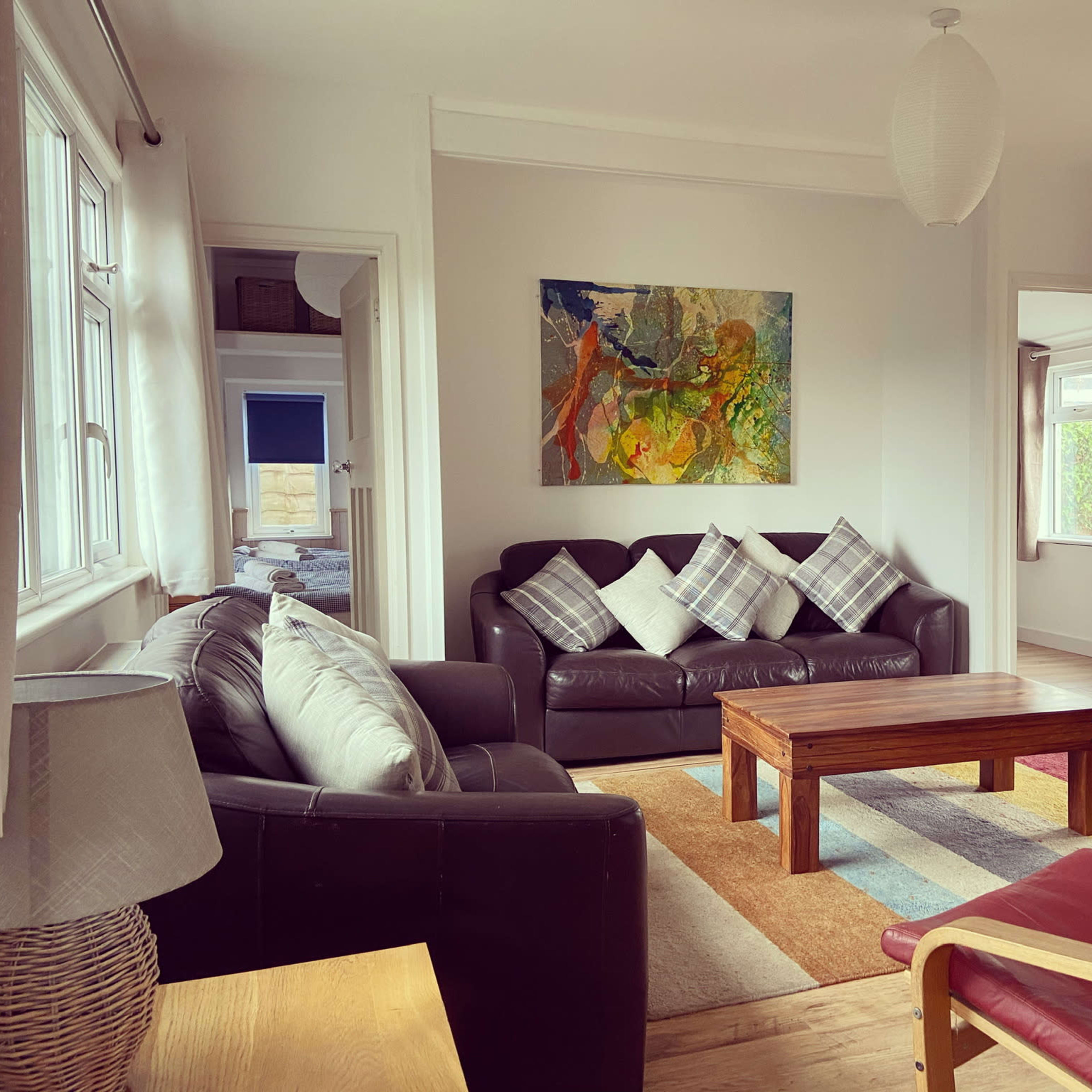 The room features a black leather sofa with plaid throw pillows, a wooden coffee table, and an abstract painting on the wall, with large windows letting in natural light.