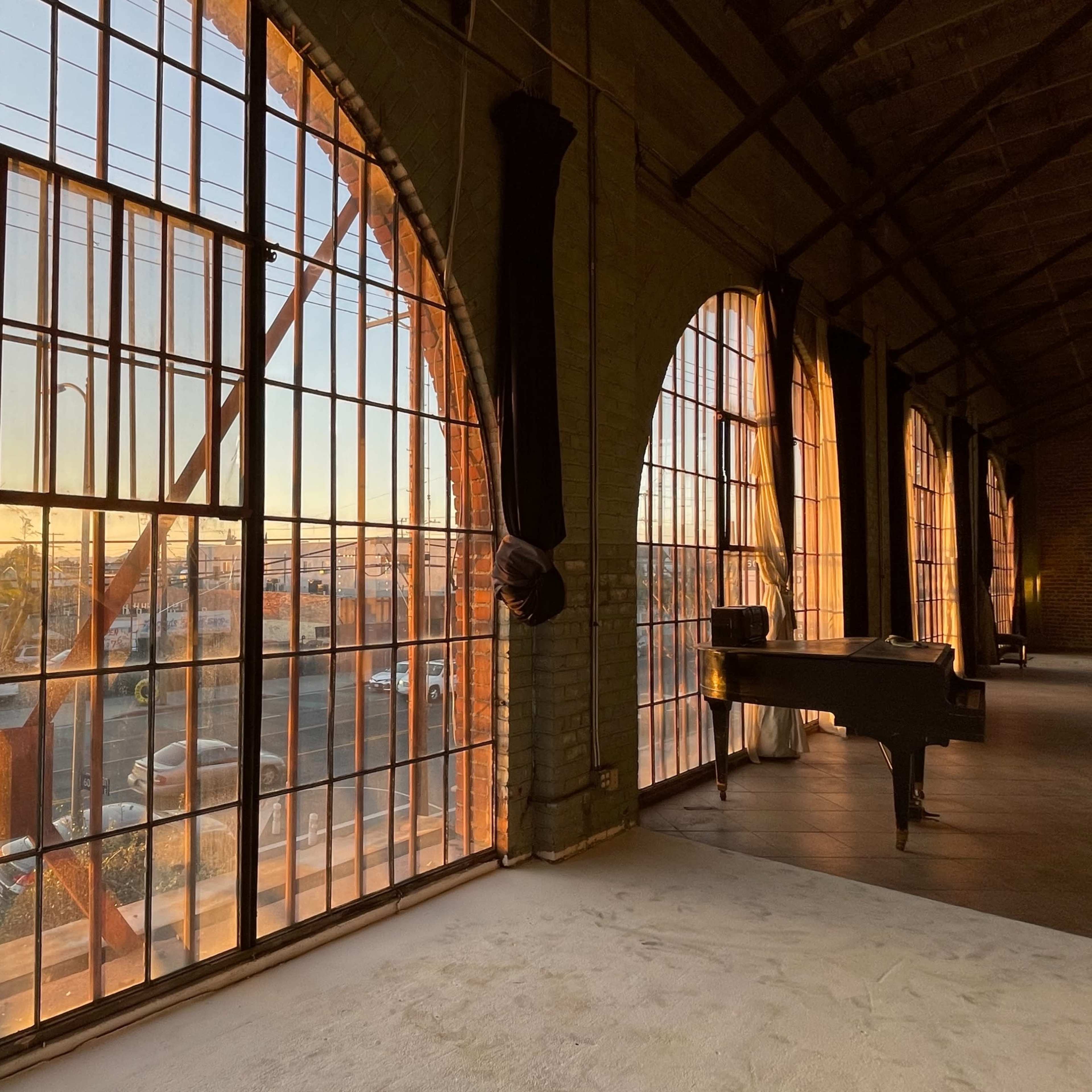 A spacious room features large industrial windows that frame a sunset view, with a piano placed near the glass.