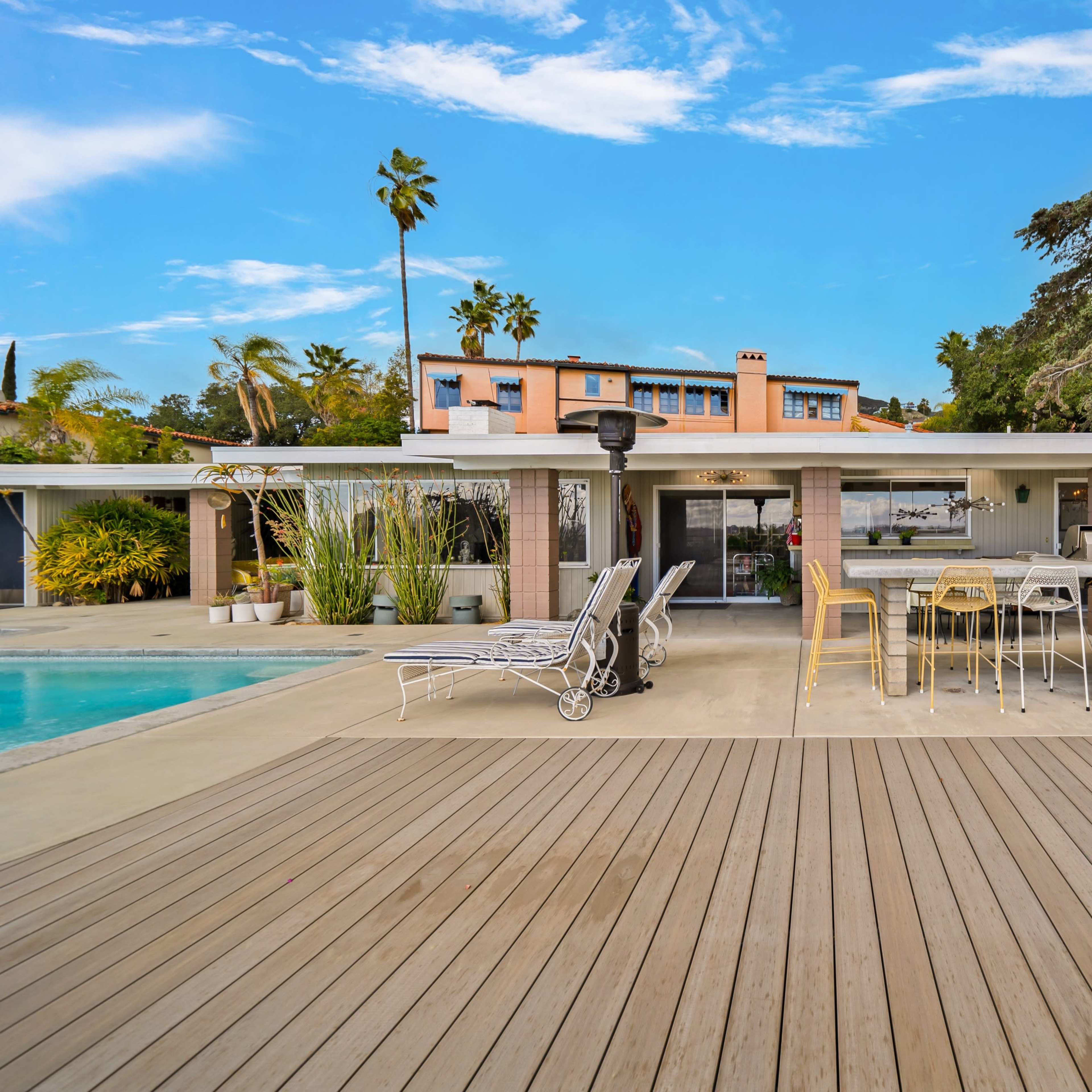 A modern outdoor patio area with a swimming pool, surrounded by lounge chairs, a bar table, and a backdrop of a residential building and palm trees.