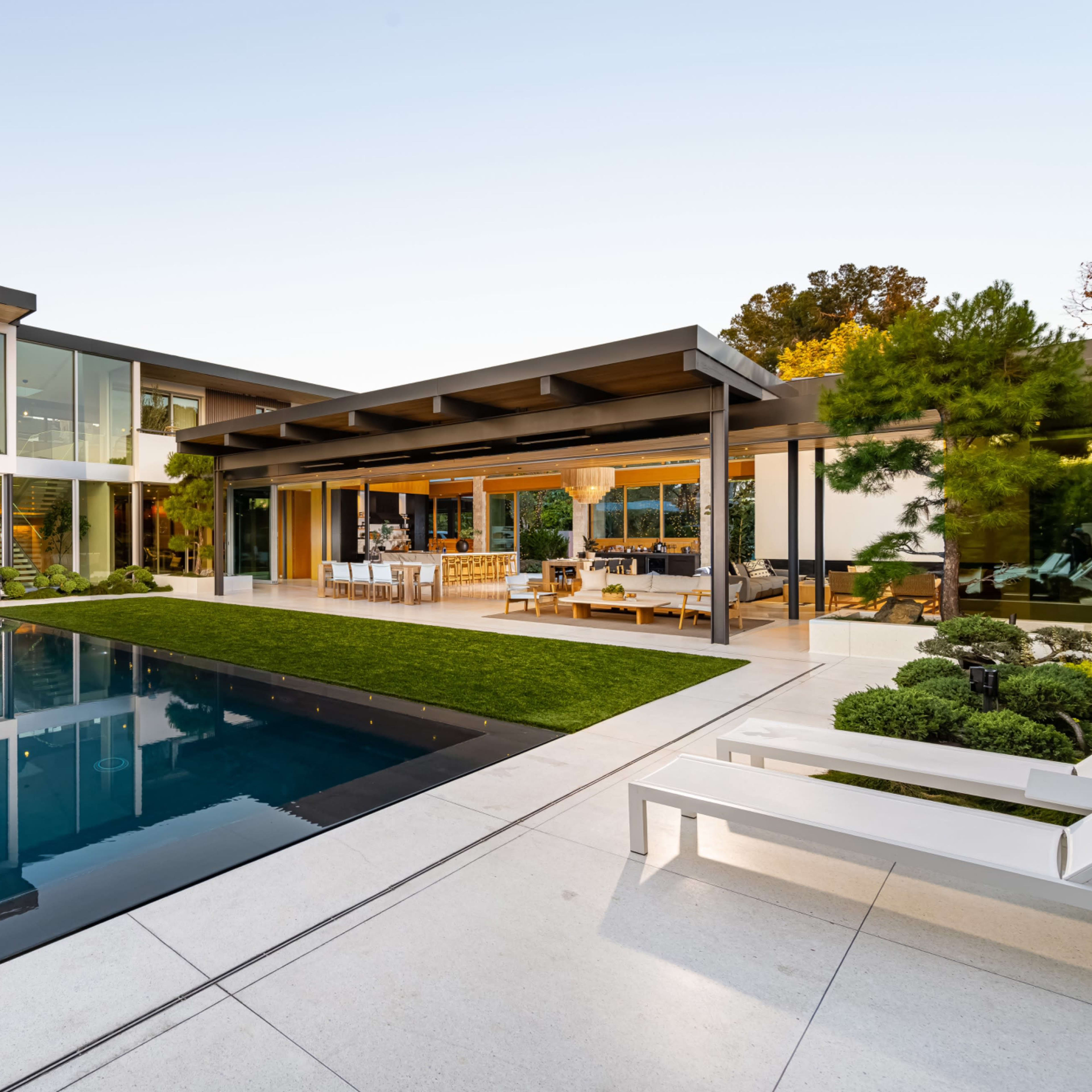 A modern home features a sleek pool surrounded by landscaped greenery and a covered outdoor seating area.