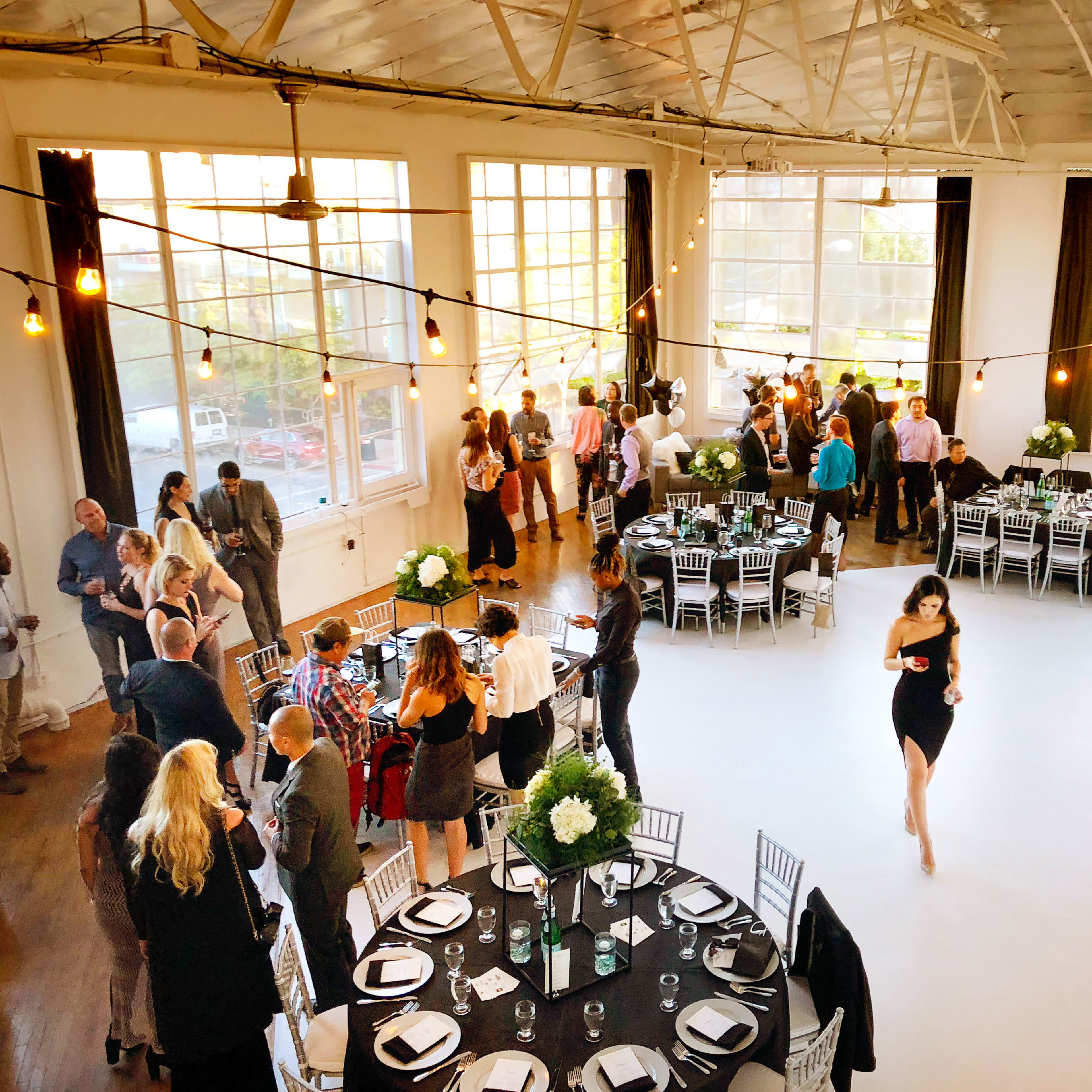 A spacious event venue is filled with people socializing at elegantly set tables, while a woman in a black dress walks across the floor.