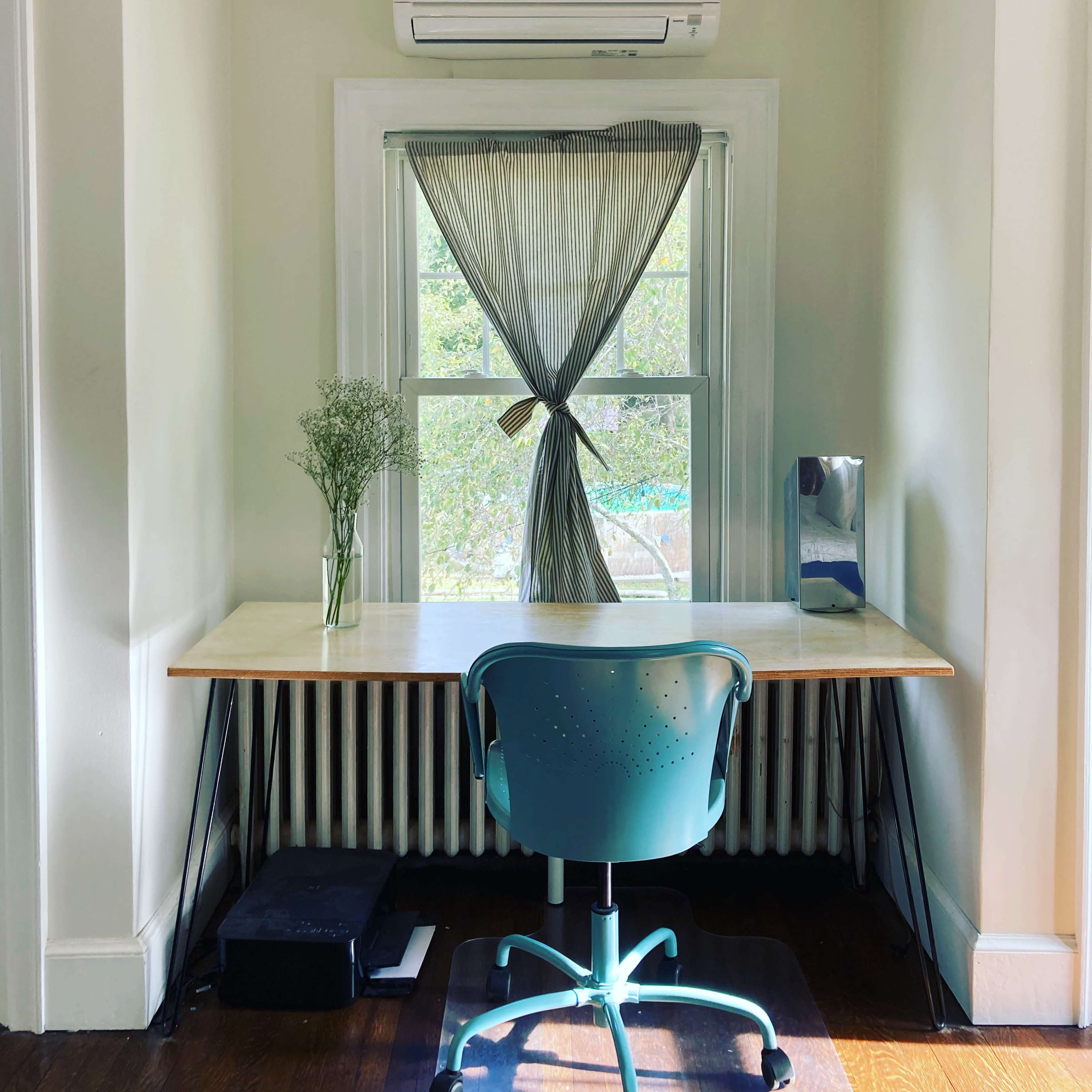 A minimalist home office features a simple desk with a chair, a vase of flowers on the desk, and a window with striped curtains above a radiator.