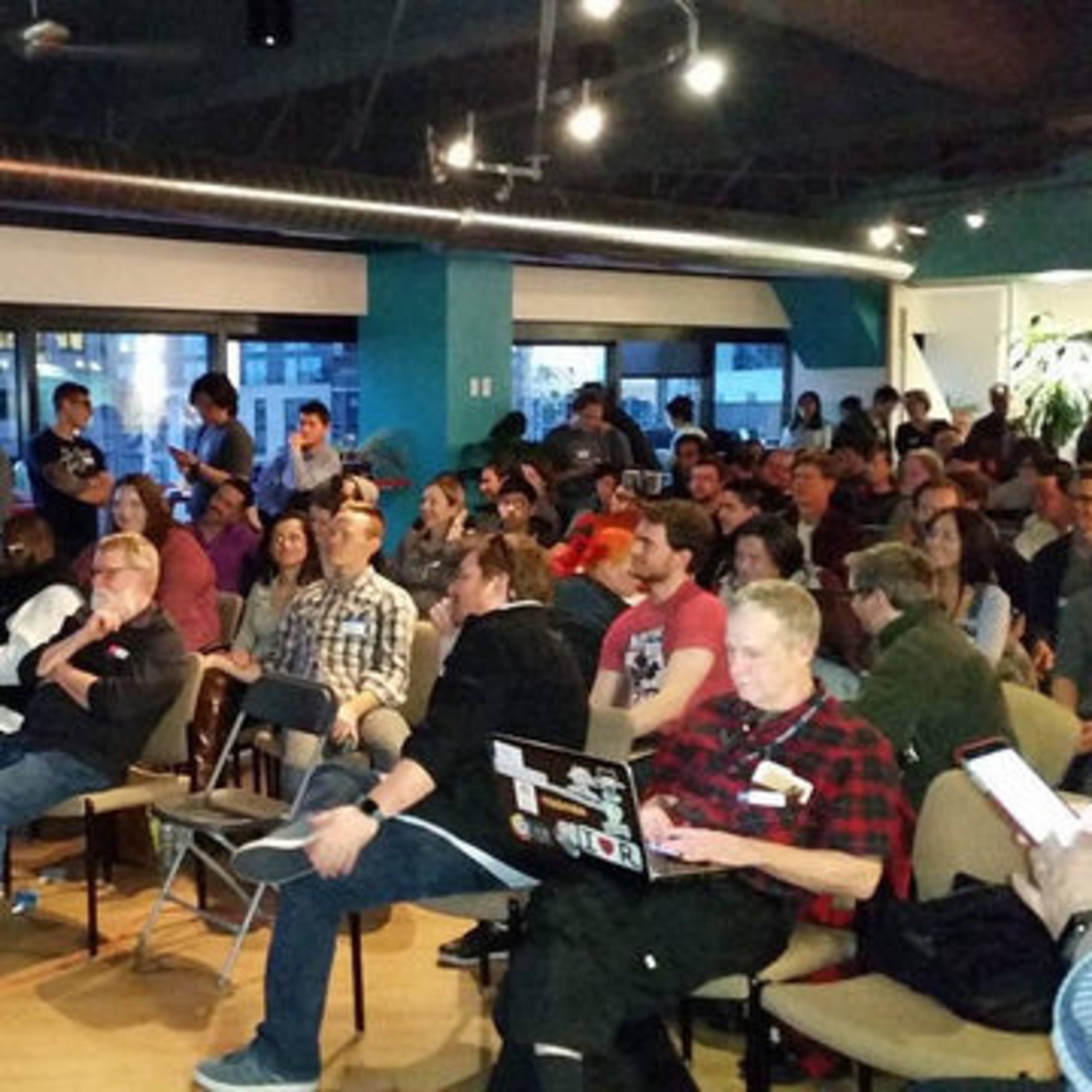 A large group of people is seated in a modern room, attentively watching a speaker at the front of the space.
