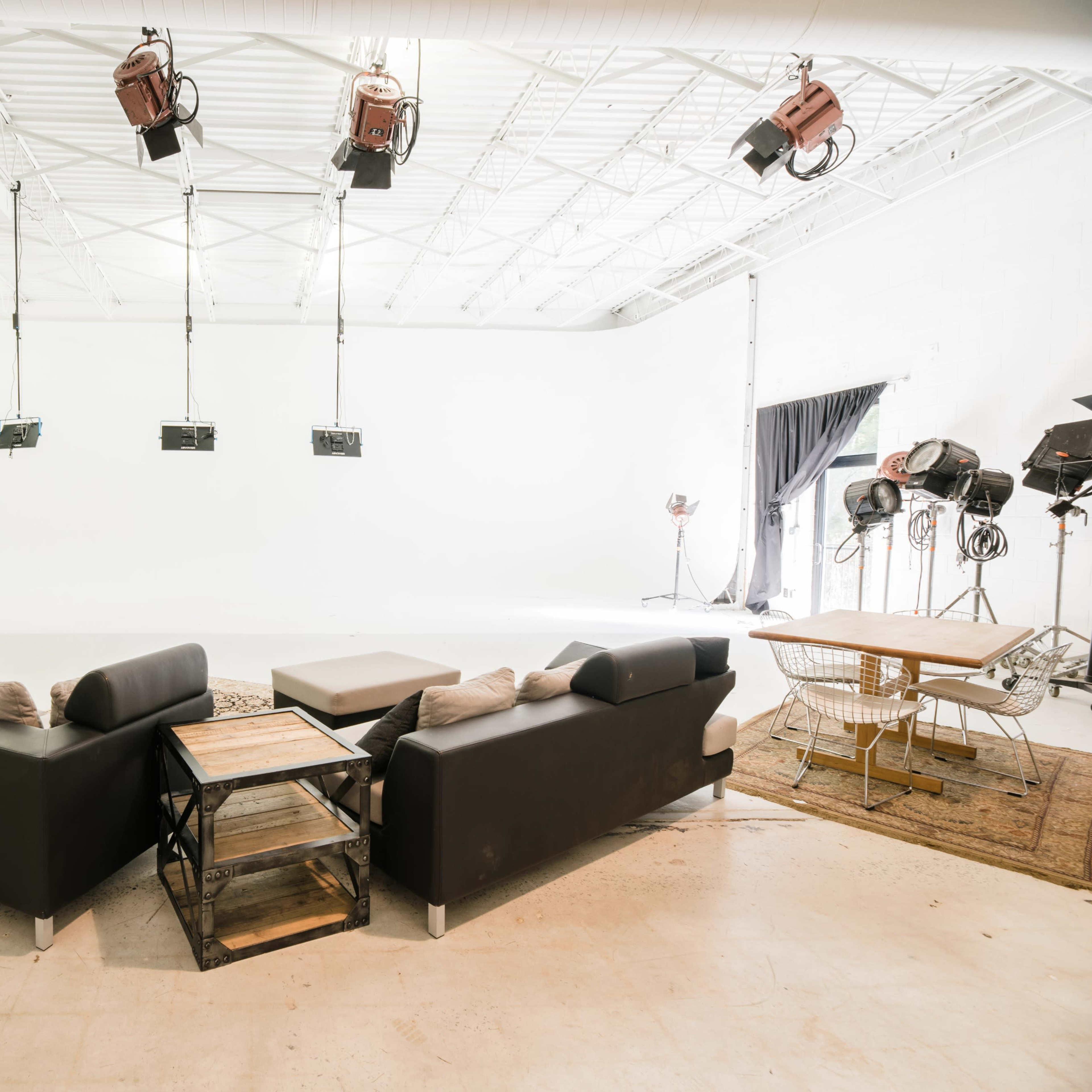 A spacious indoor studio with a black sectional sofa, a wooden coffee table, and several studio lights overhead.