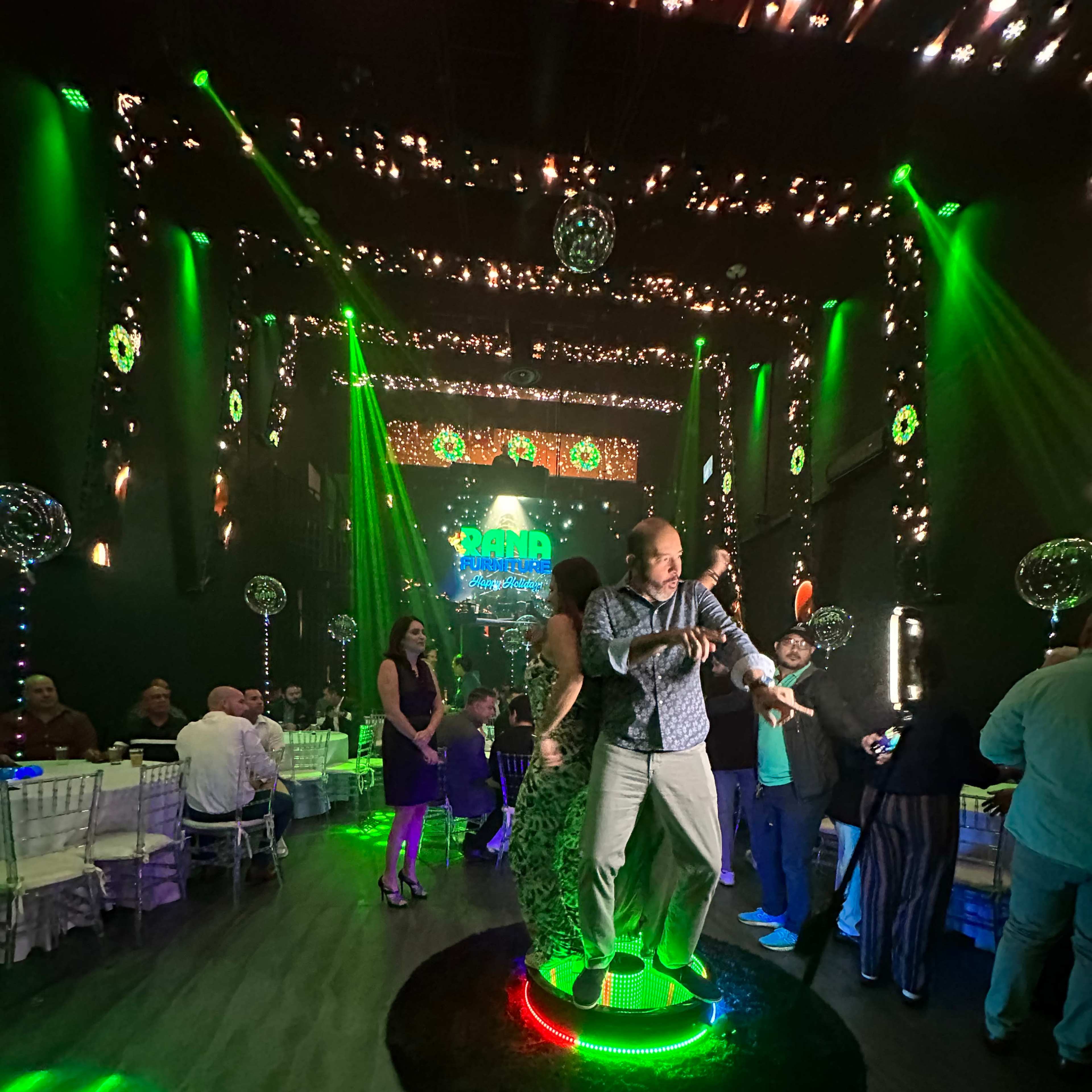 A man lifts a woman while dancing on a circular illuminated platform in a festive venue decorated with green lights and sparkles.