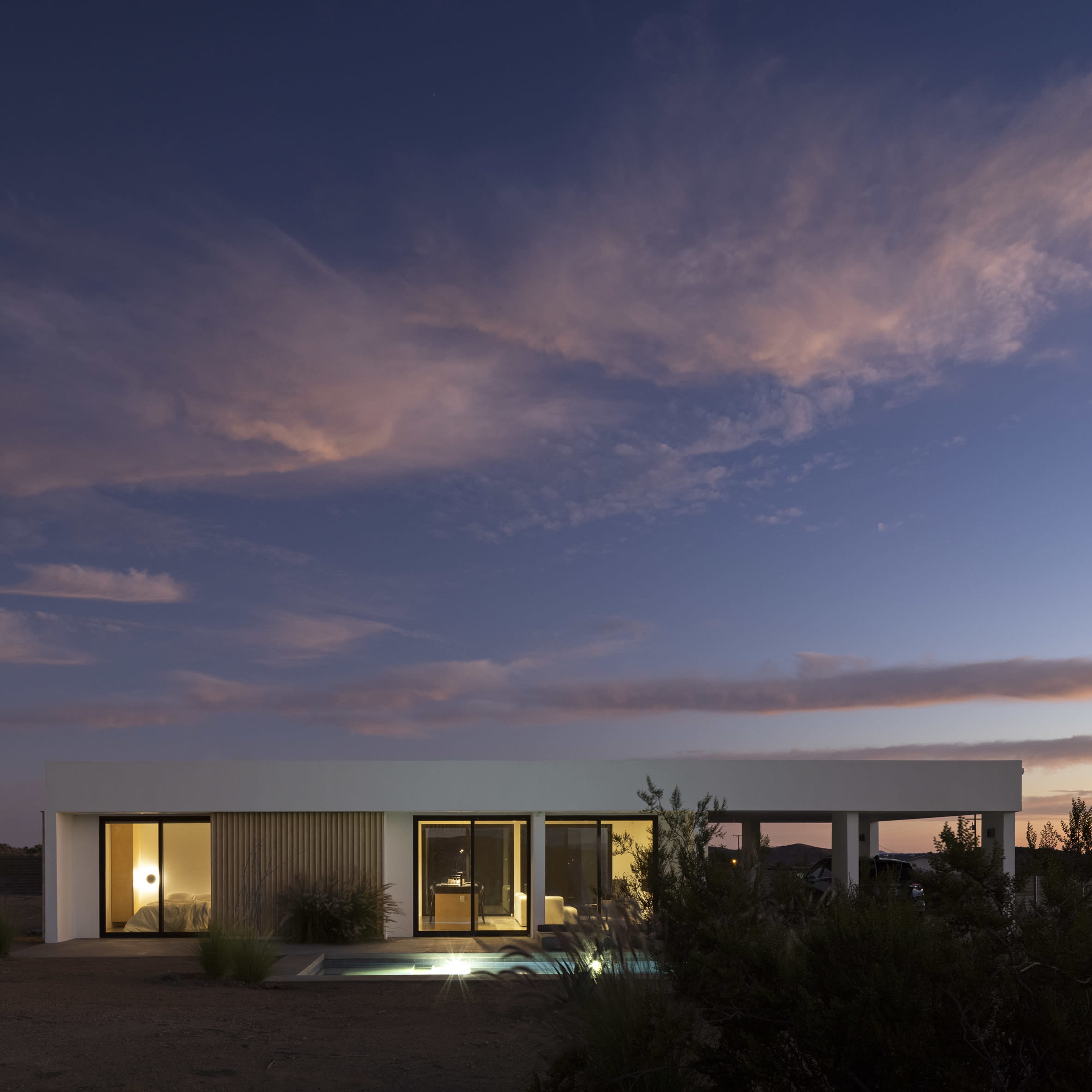 A modern, single-story house with large windows and a minimalistic design sits in a desert landscape at dusk, with soft clouds in the sky.