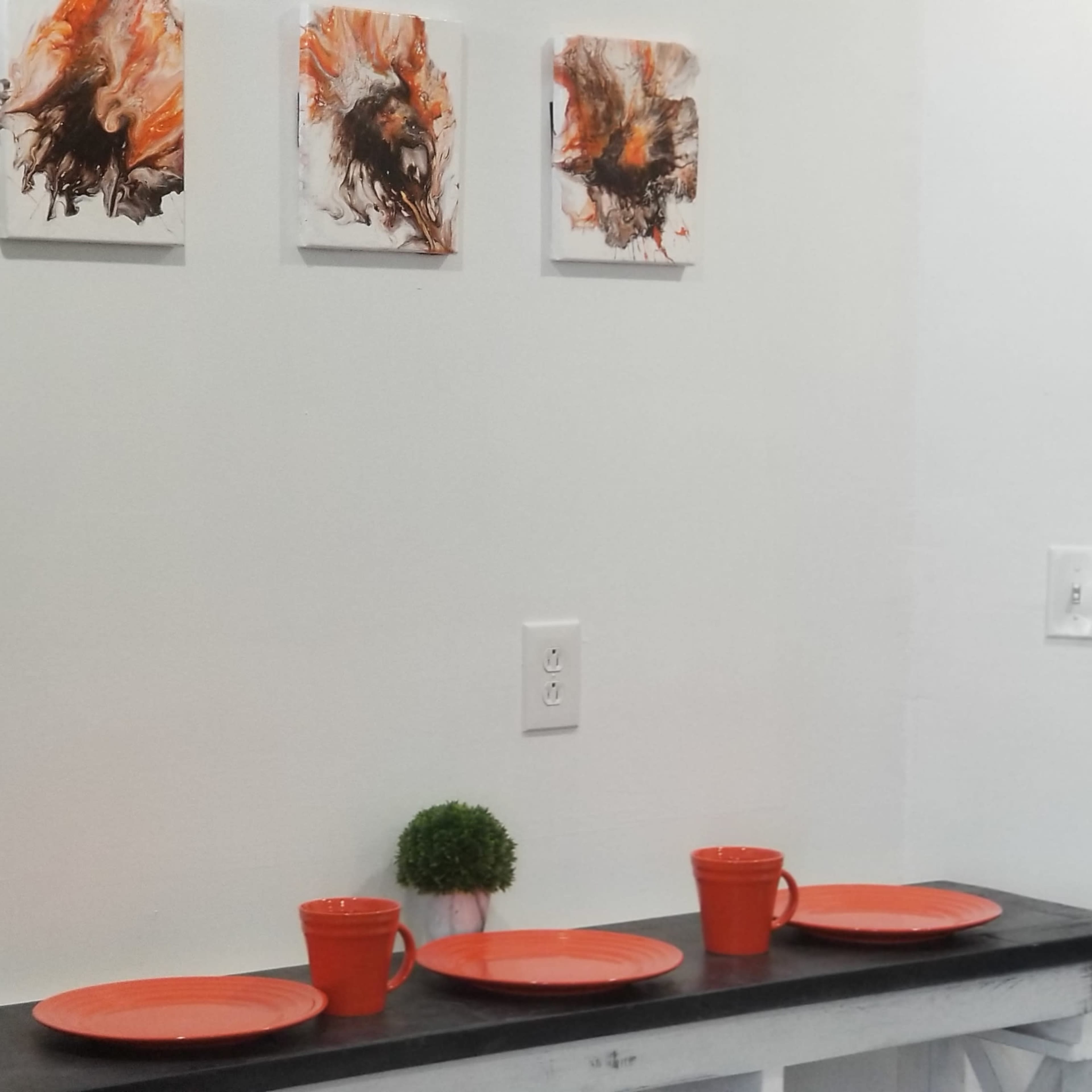 A white wall with three abstract art pieces in orange and black above a black countertop featuring four orange plates and two small orange pots with plants.